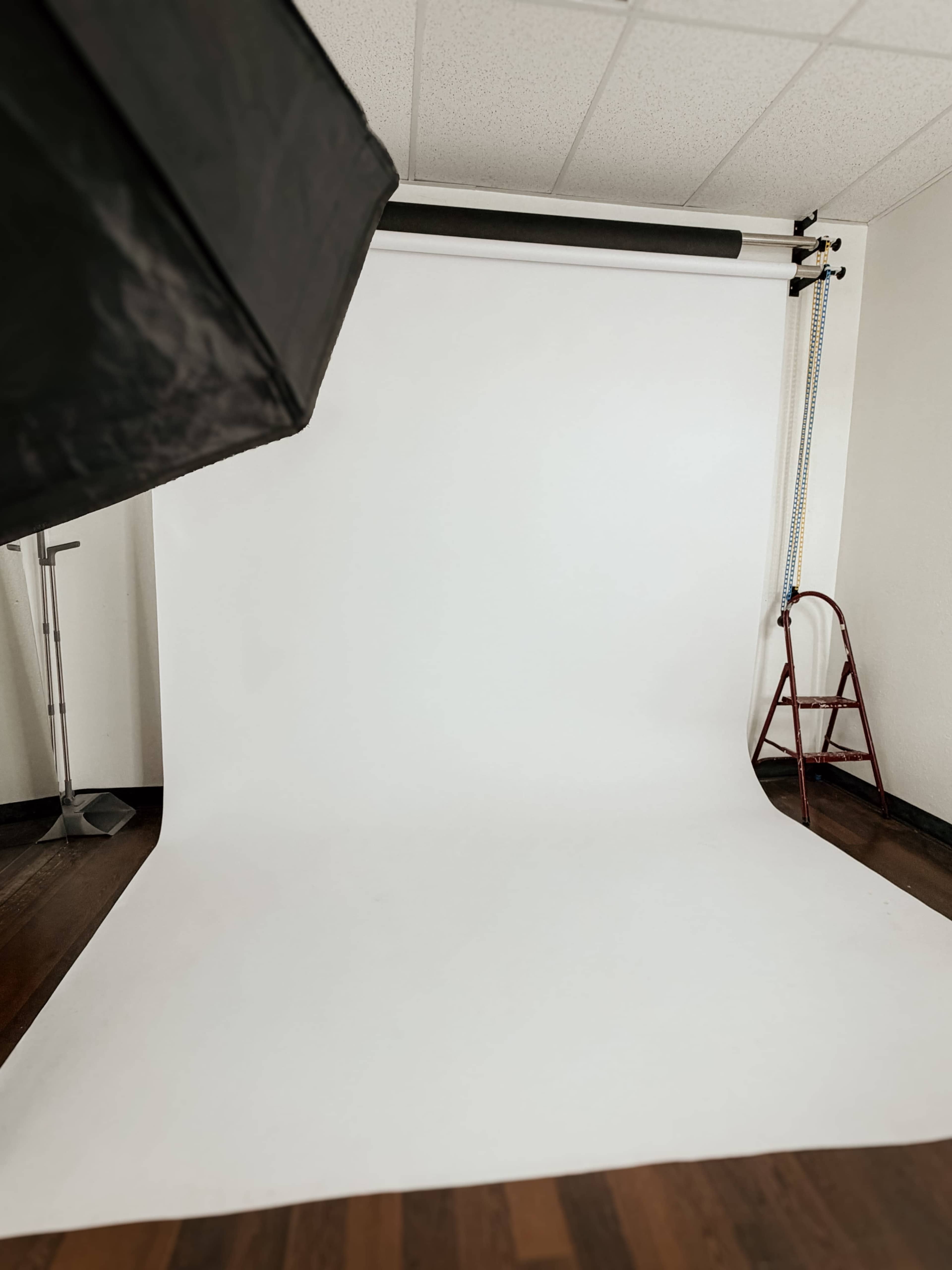 The image depicts a photography studio setup featuring a white backdrop, a ladder, and studio lighting equipment.