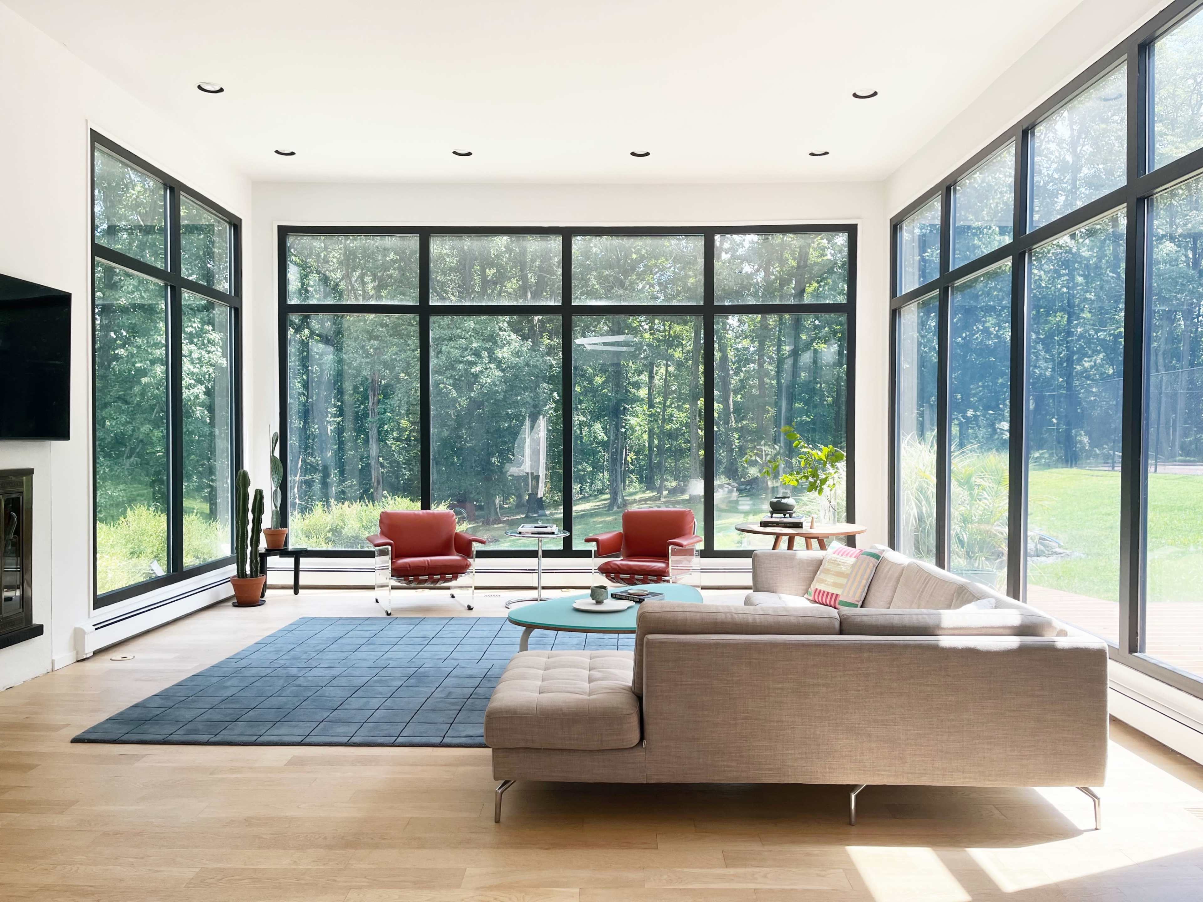 The image shows a modern living room with large windows overlooking a green outdoor space, featuring a sectional gray sofa, red armchairs, and a blue coffee table on a dark rug.