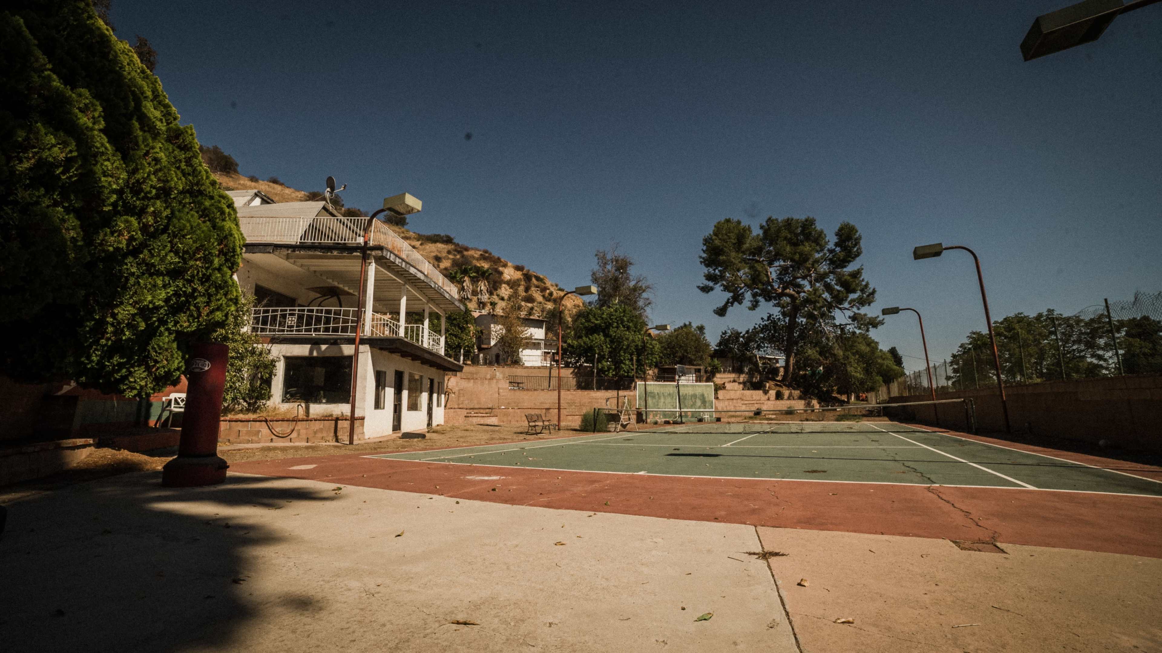 A vacant tennis court surrounded by a two-story building and trees, set against a hilly background under a clear sky.