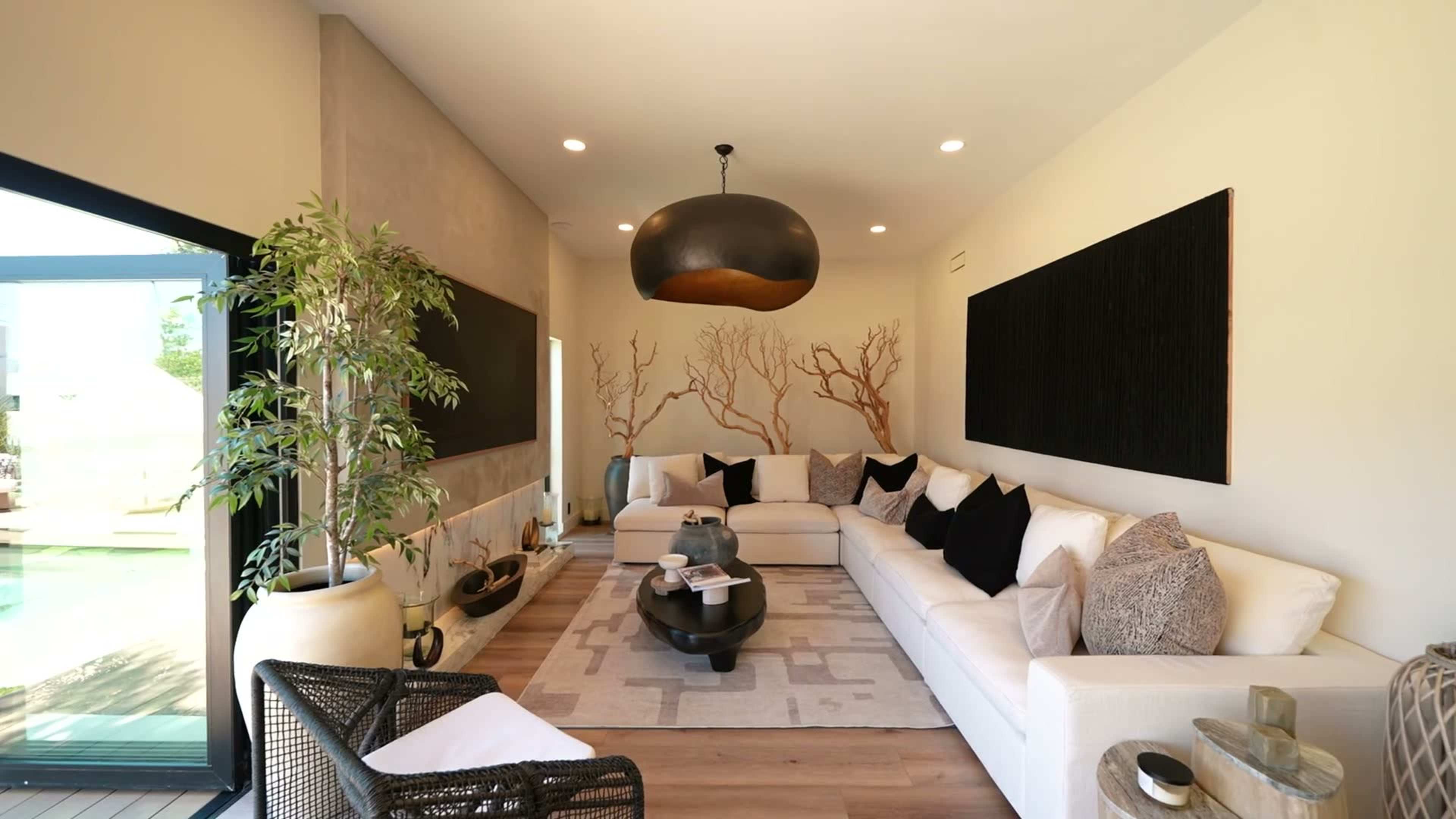 The image shows a modern living room featuring a large white sectional sofa, a round black coffee table, and decorative wall art with tree patterns, complemented by potted plants and natural light from nearby windows.