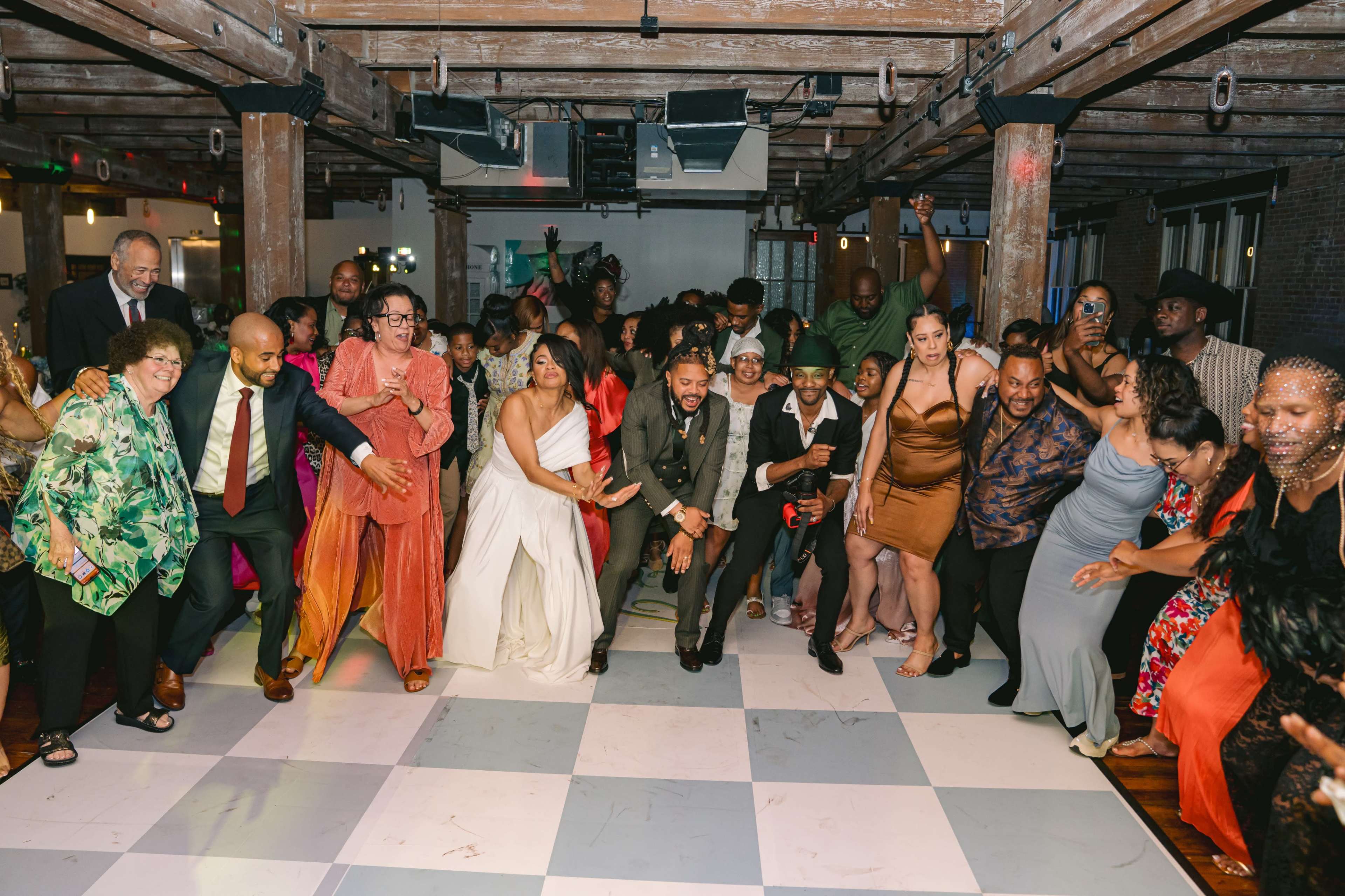 A large group of people dance joyfully at a lively indoor event with a checkered floor and wooden beams above.