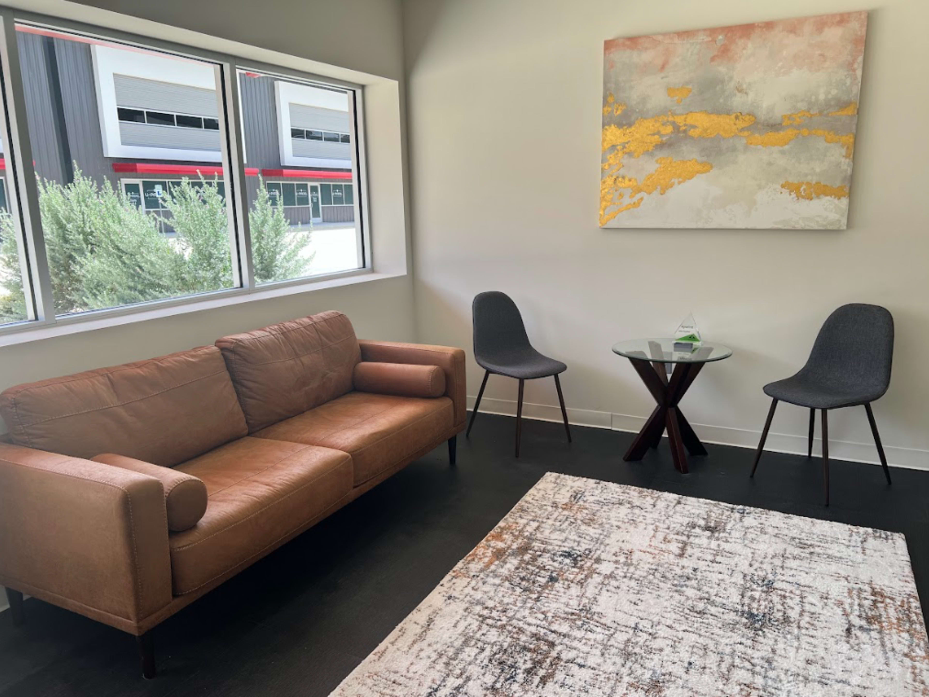 The image shows a modern waiting area featuring a brown leather sofa, two chairs, a small glass table, and an abstract painting on the wall.
