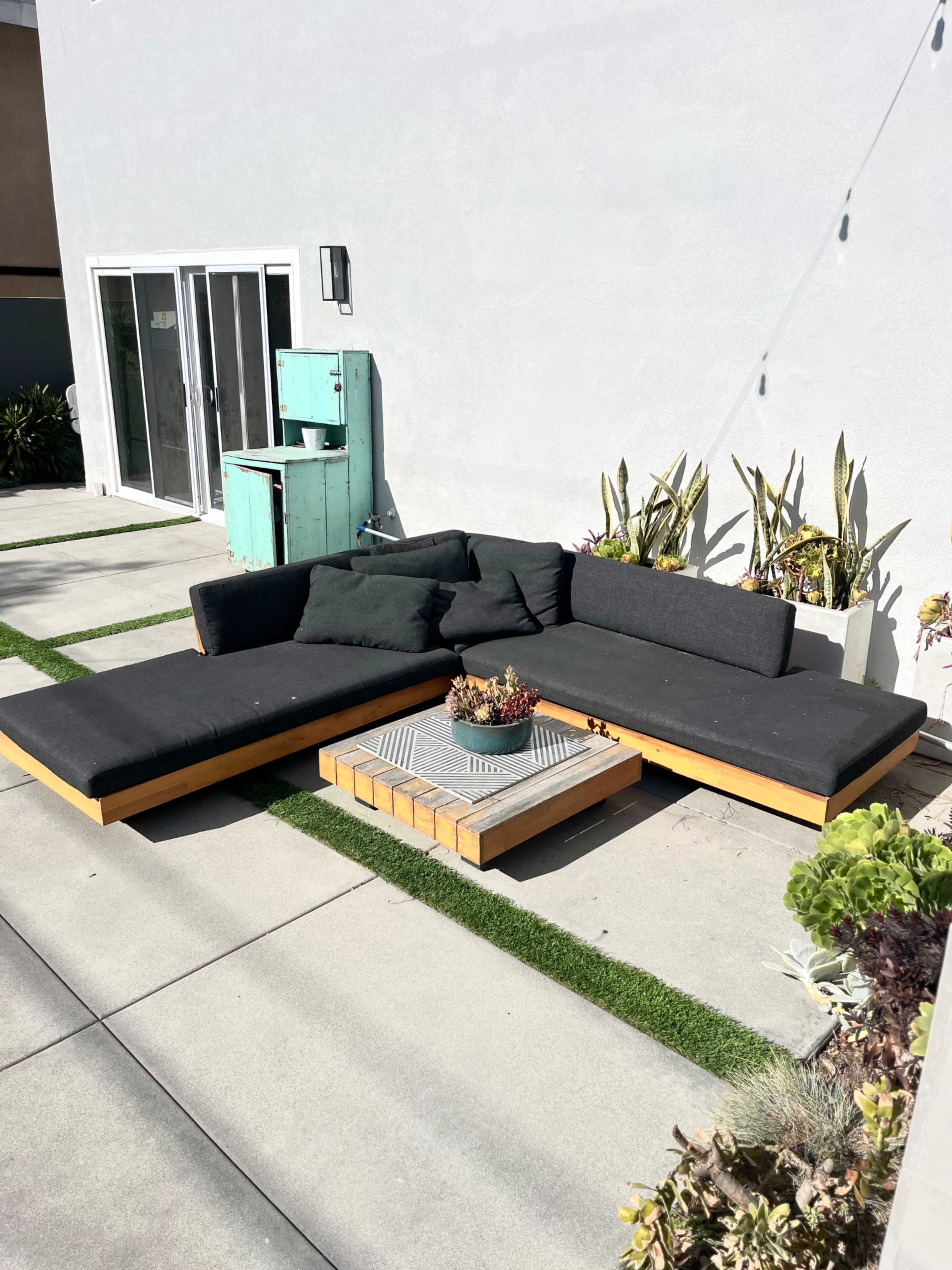 A modular outdoor sectional sofa with a wooden frame and a stone coffee table sits on a concrete patio beside a green lawn and a small turquoise cabinet.