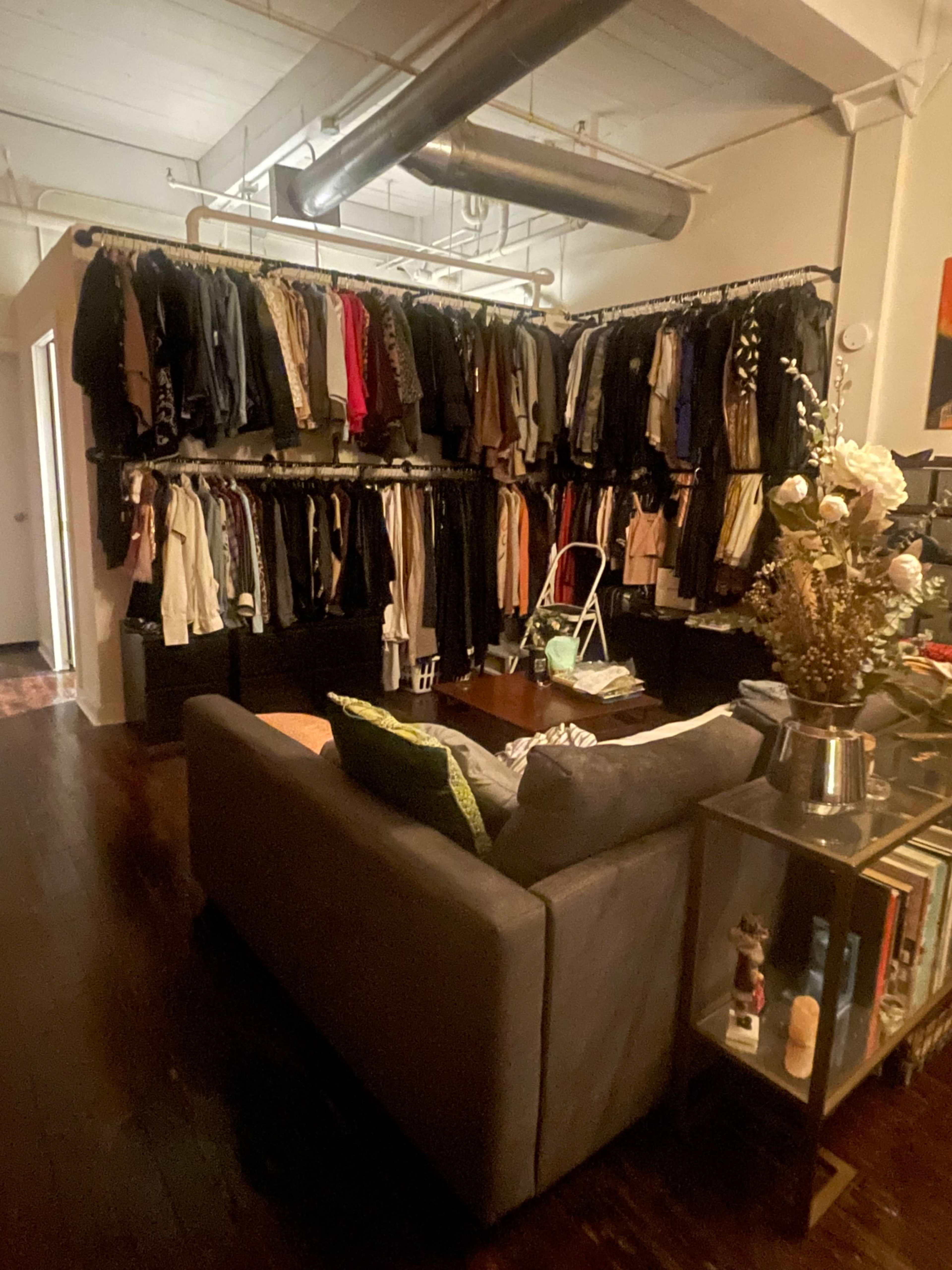 A cozy living space features a gray sofa facing a wall of organized clothing hung on racks, with a small coffee table and decorative items nearby.