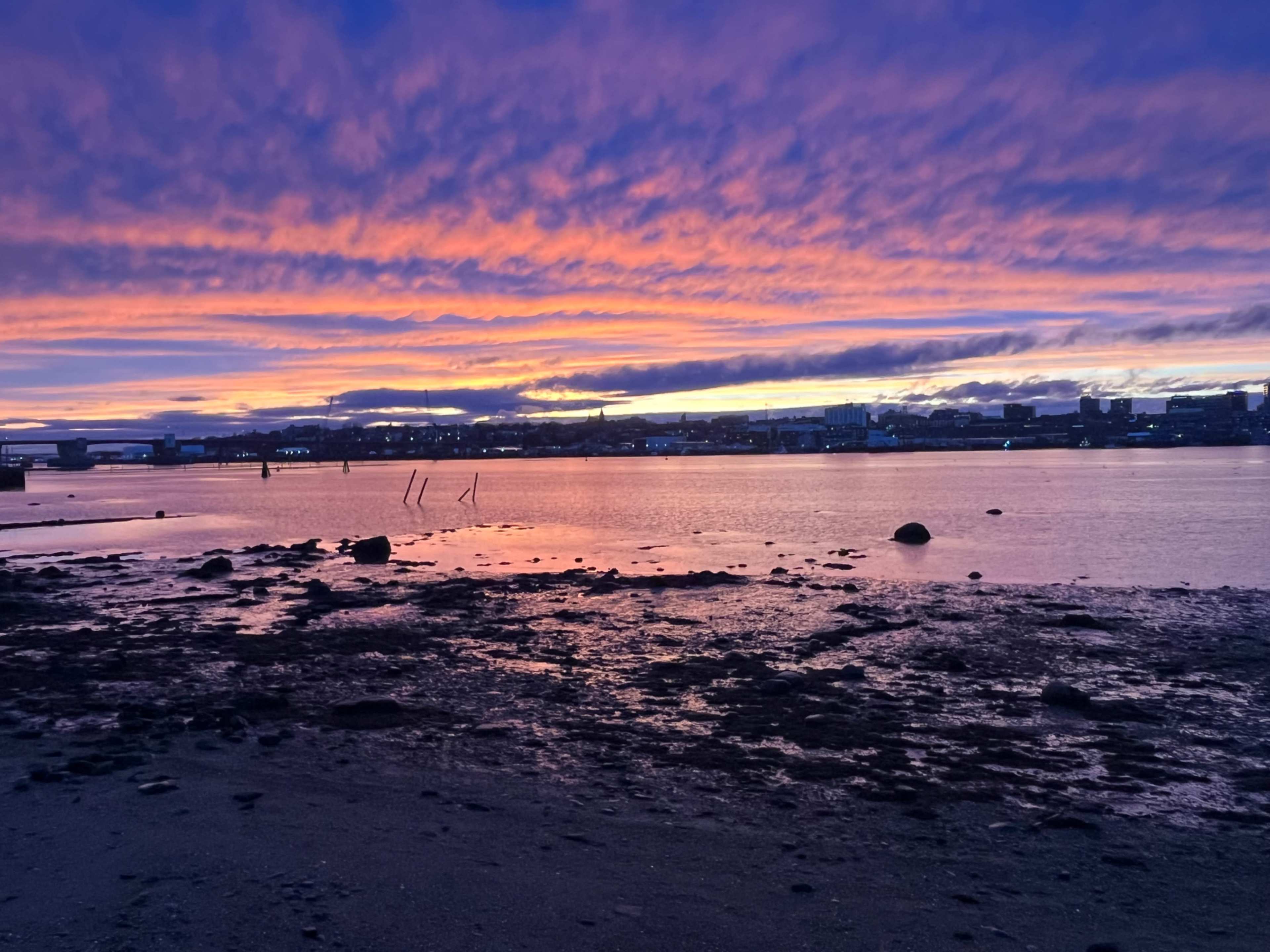 The image shows a riverbank at sunset with colorful clouds reflecting on the water and rocks scattered along the shore.