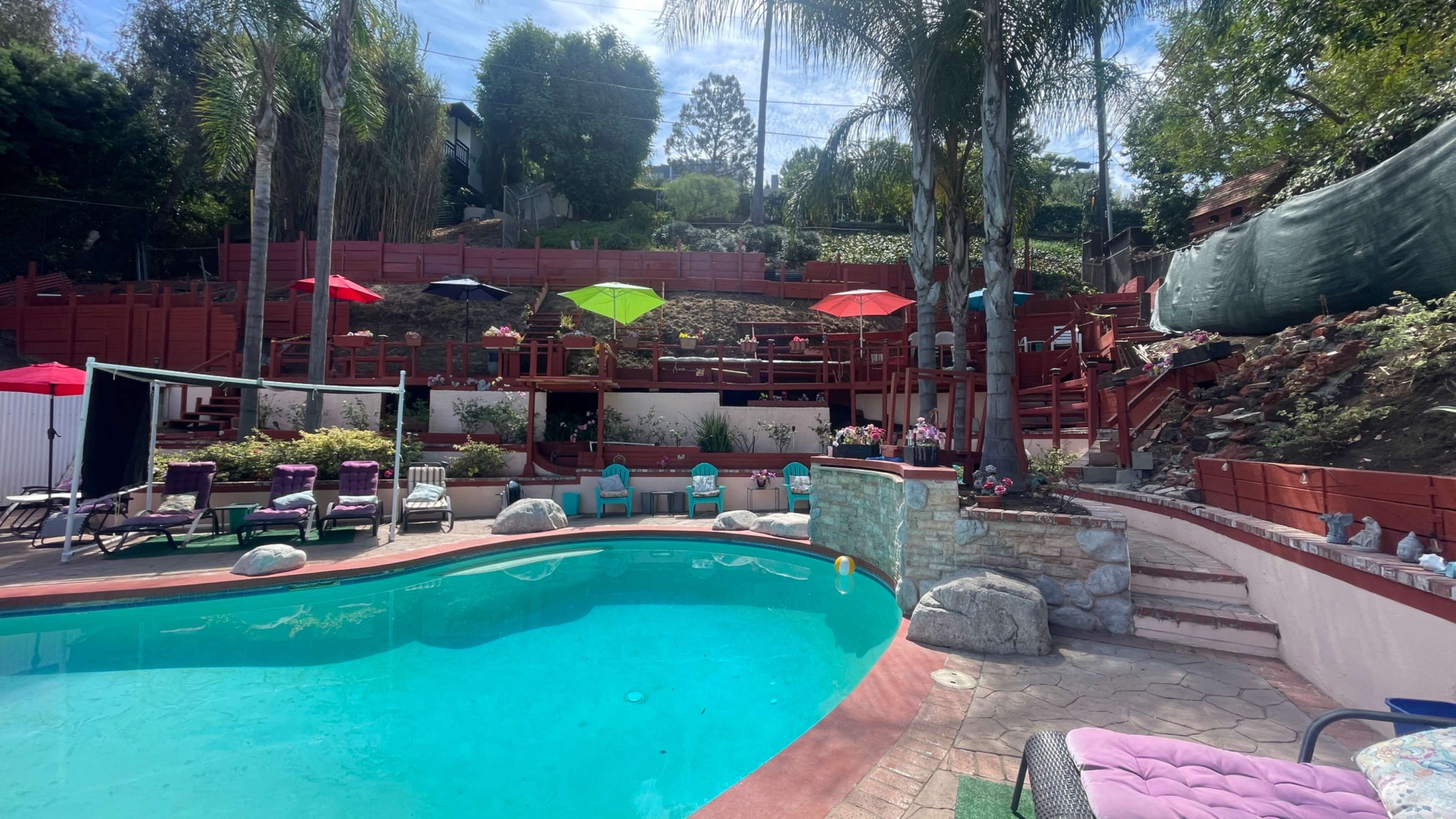 A swimming pool is surrounded by lounge chairs and umbrellas in a landscaped backyard with tiered seating and a sloped area in the background.
