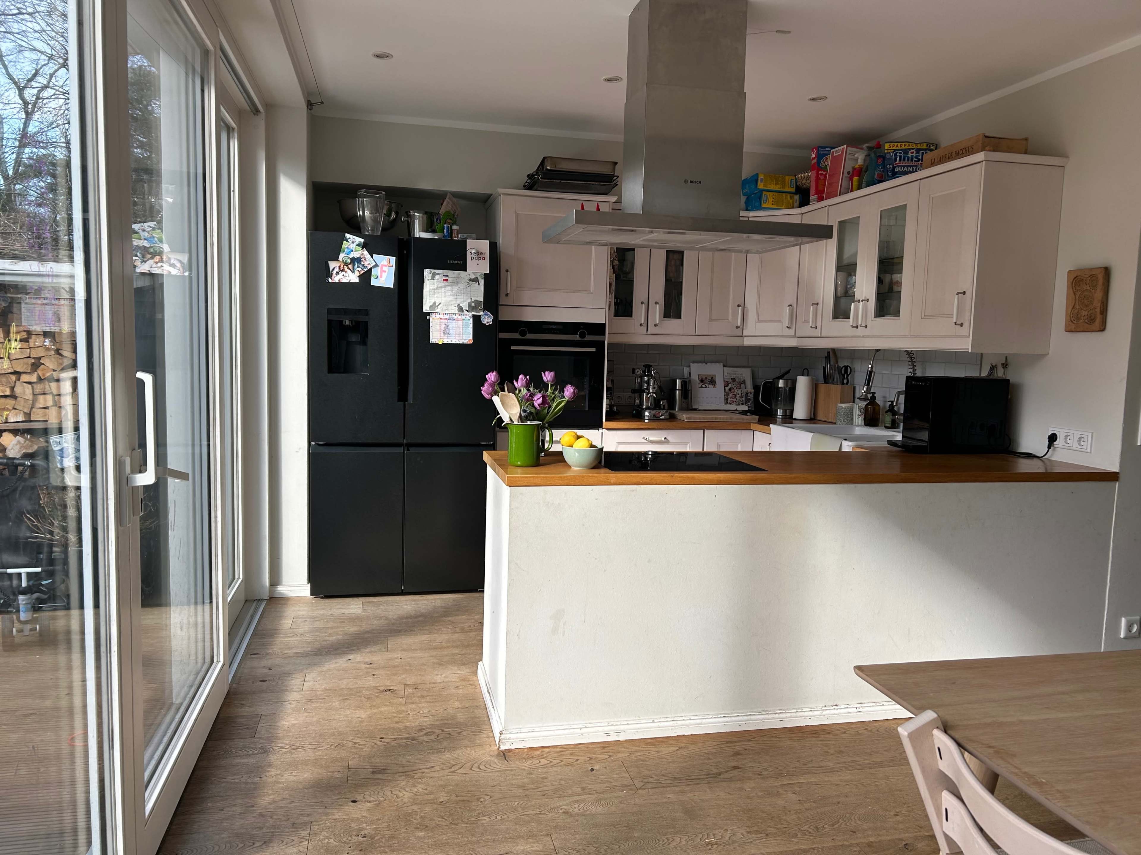 A modern kitchen features a central island with a stovetop, white cabinetry, and a black refrigerator, illuminated by natural light from large windows.