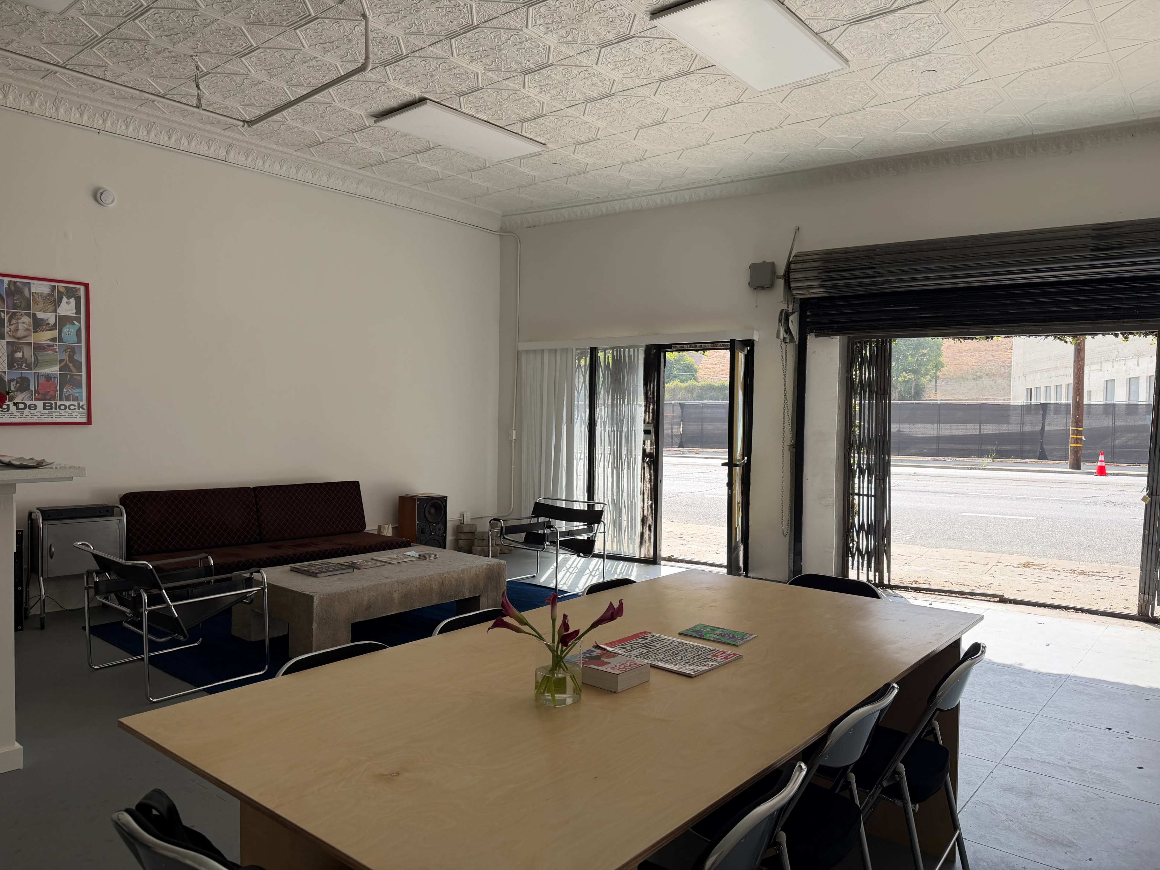 The image shows a bright indoor space featuring a large wooden table surrounded by chairs, a sofa along one wall, and large windows with doors that open to the street.