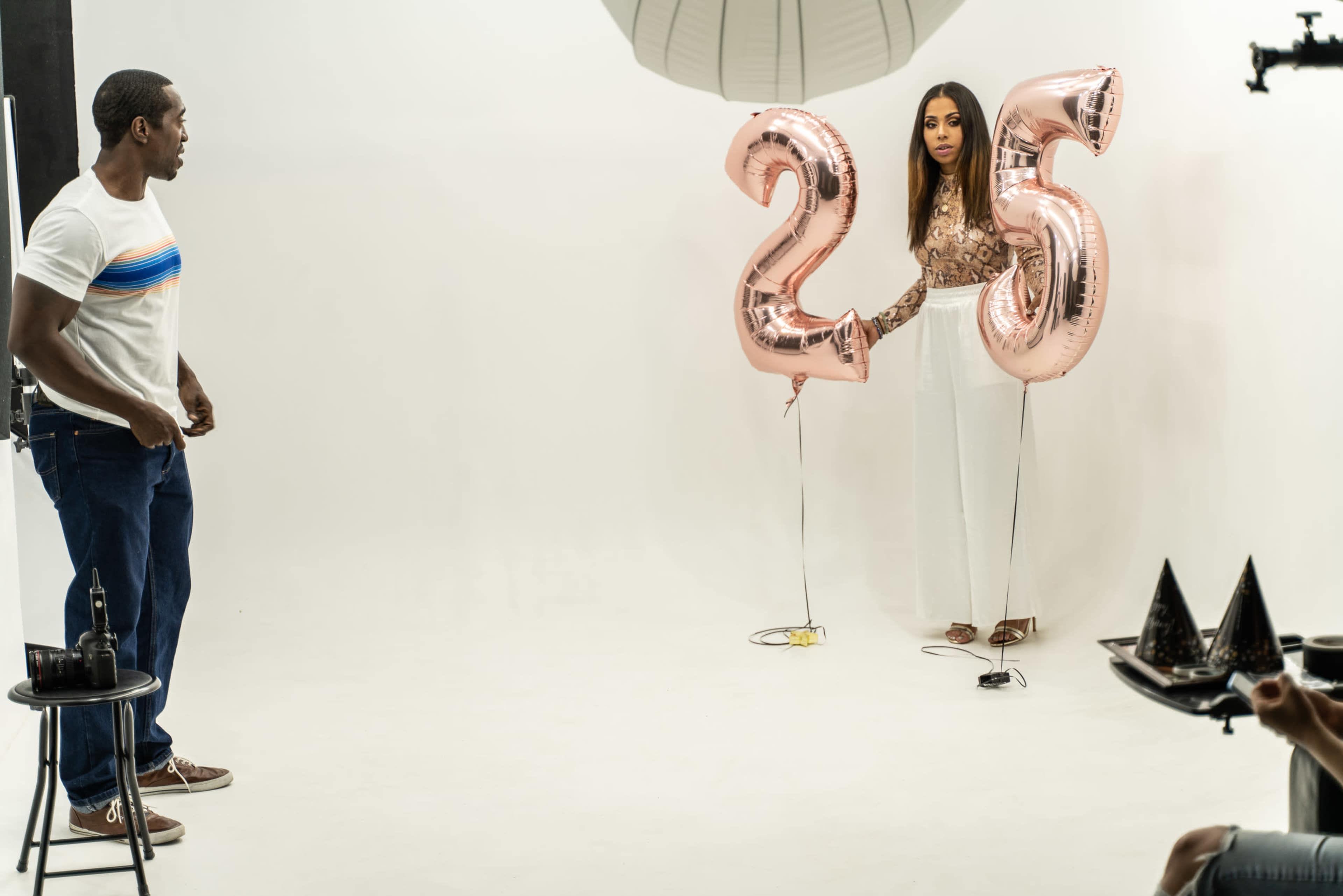 A man stands on one side of a studio while a woman holds two large rose gold balloons that read "25" on the other side.