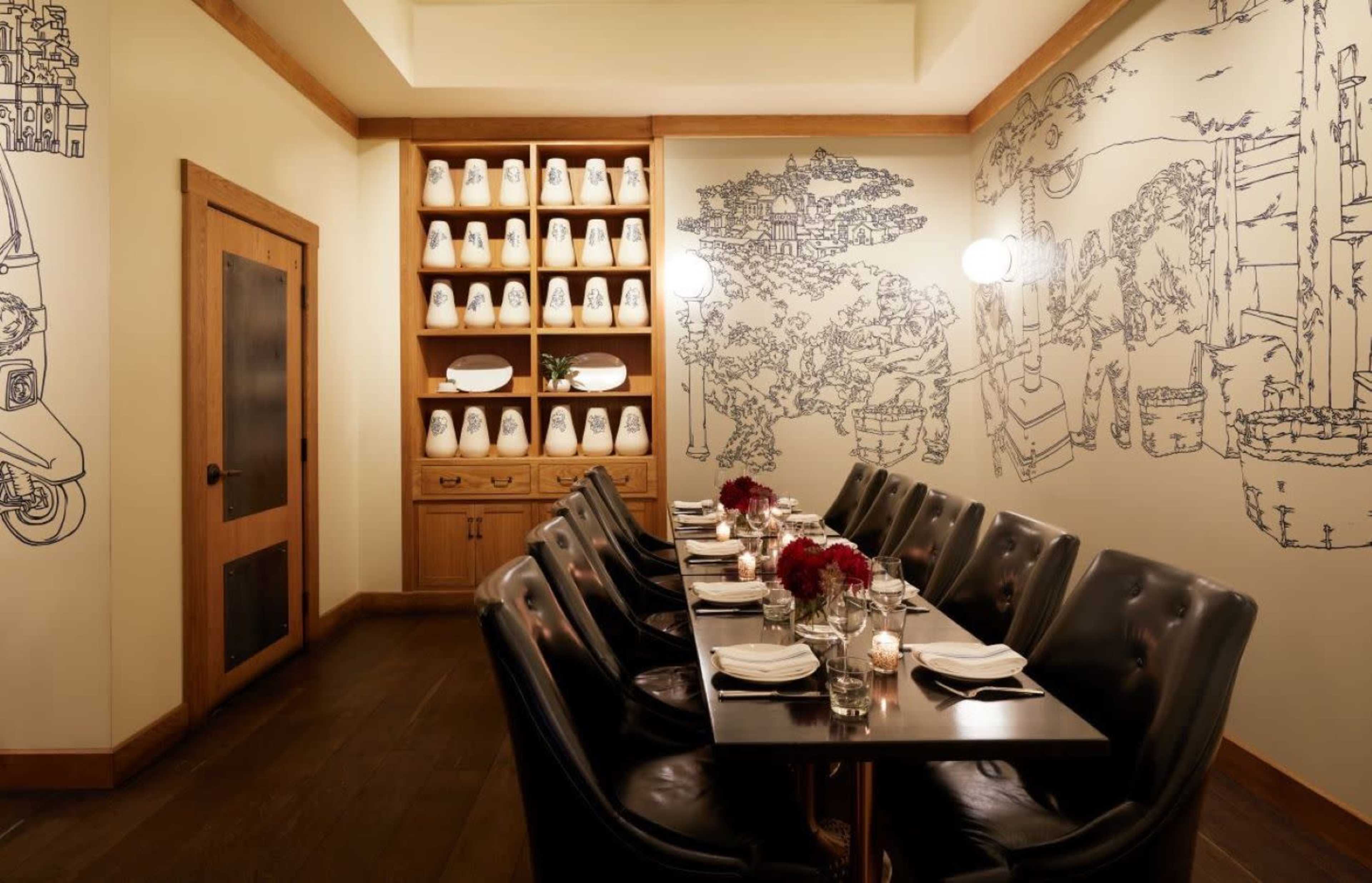 A long, dark wooden table is set for a meal in a restaurant, surrounded by black leather chairs, with minimalist line-drawn décor on the walls and a shelf displaying white dishes in the background.