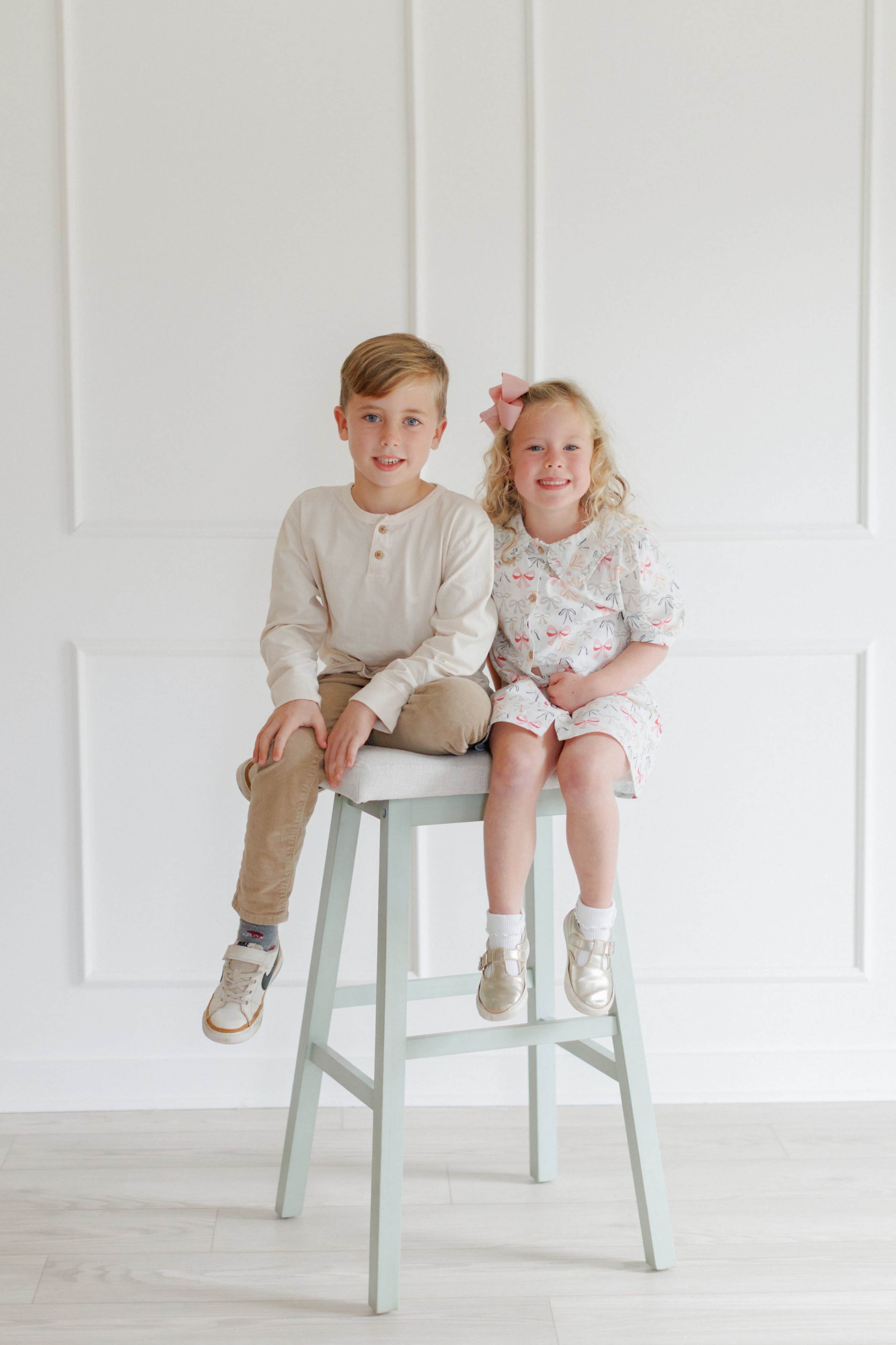 Two young children, a boy and a girl, sit on a light blue stool against a plain white wall.