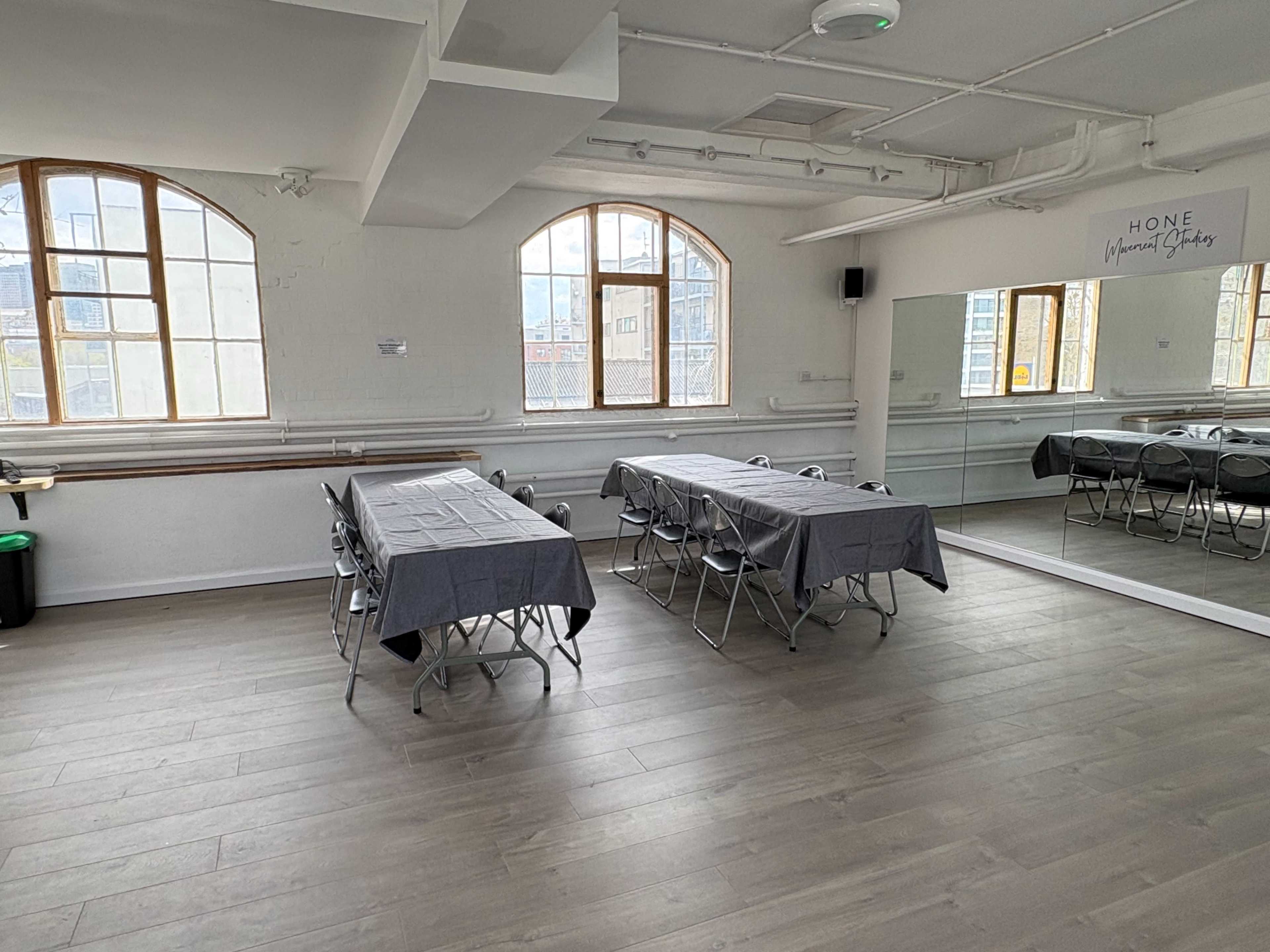 The image shows a bright, minimalist room featuring two tables covered with gray tablecloths and surrounded by folding chairs, with large windows and a mirror on one wall.