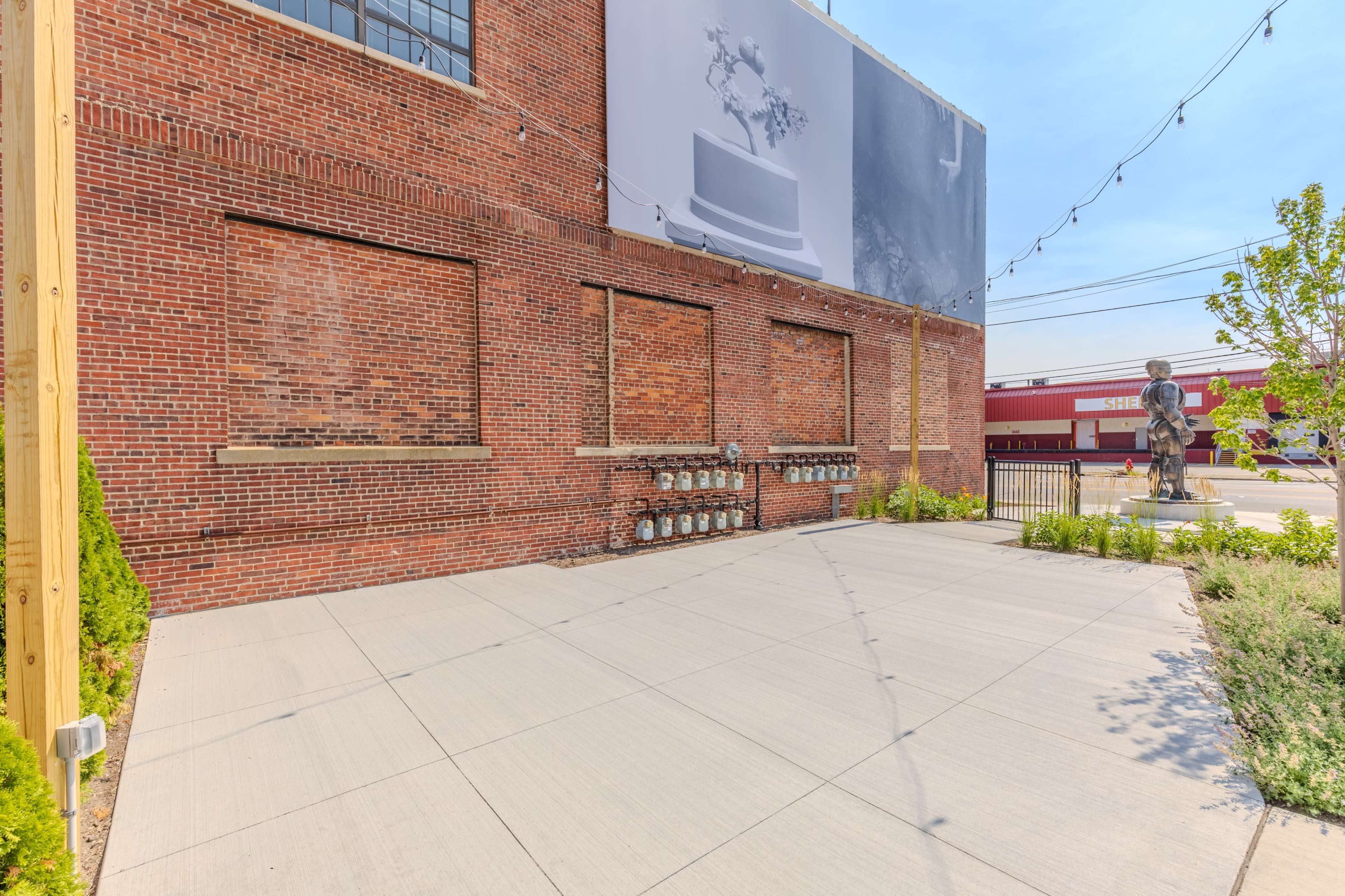The image shows a brick wall with several empty window openings, a large banner above, and a paved area in front featuring greenery and a statue.