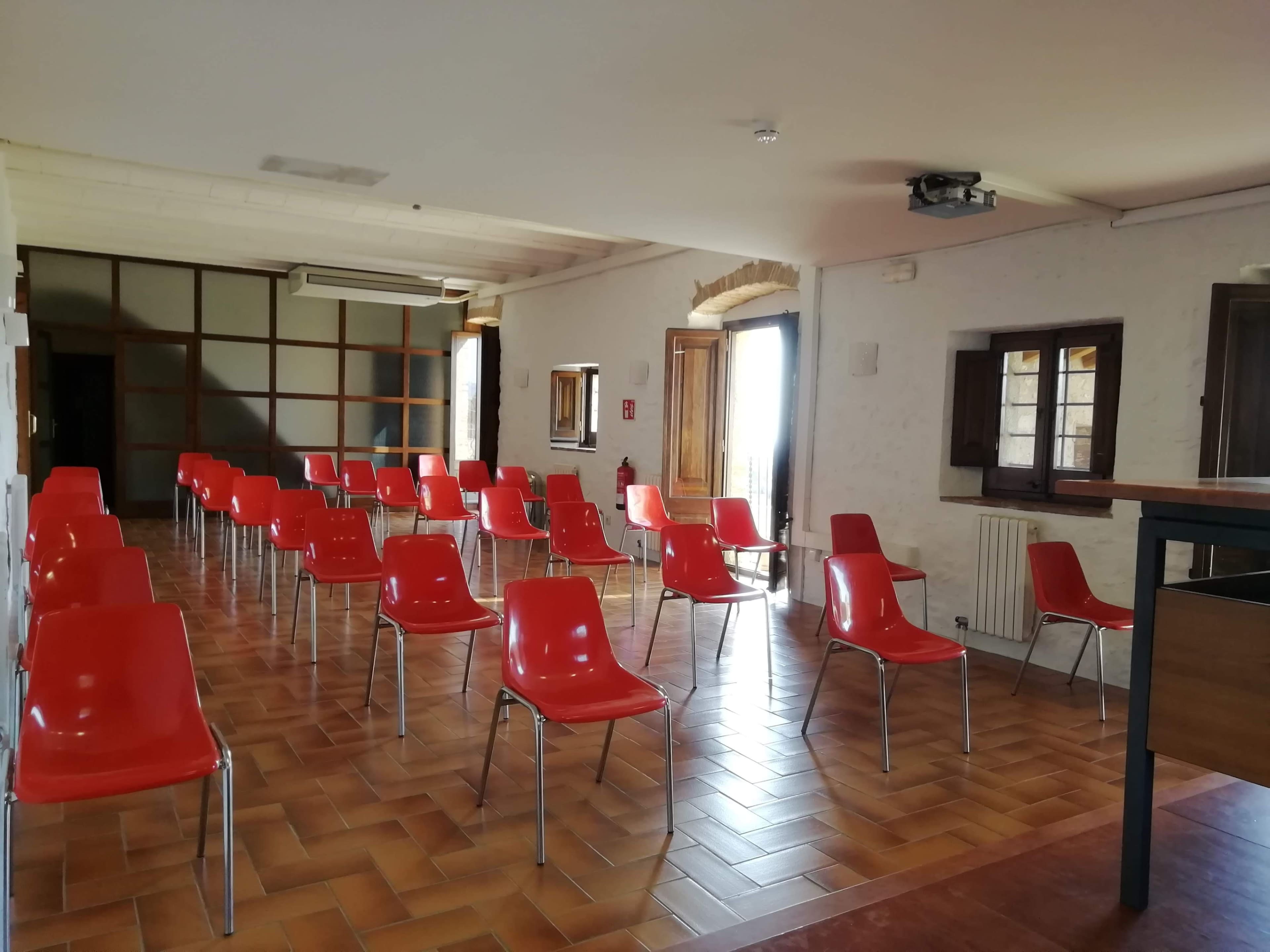 The image shows a room arranged with multiple red chairs facing a projection area, set on a tiled floor with windows around.