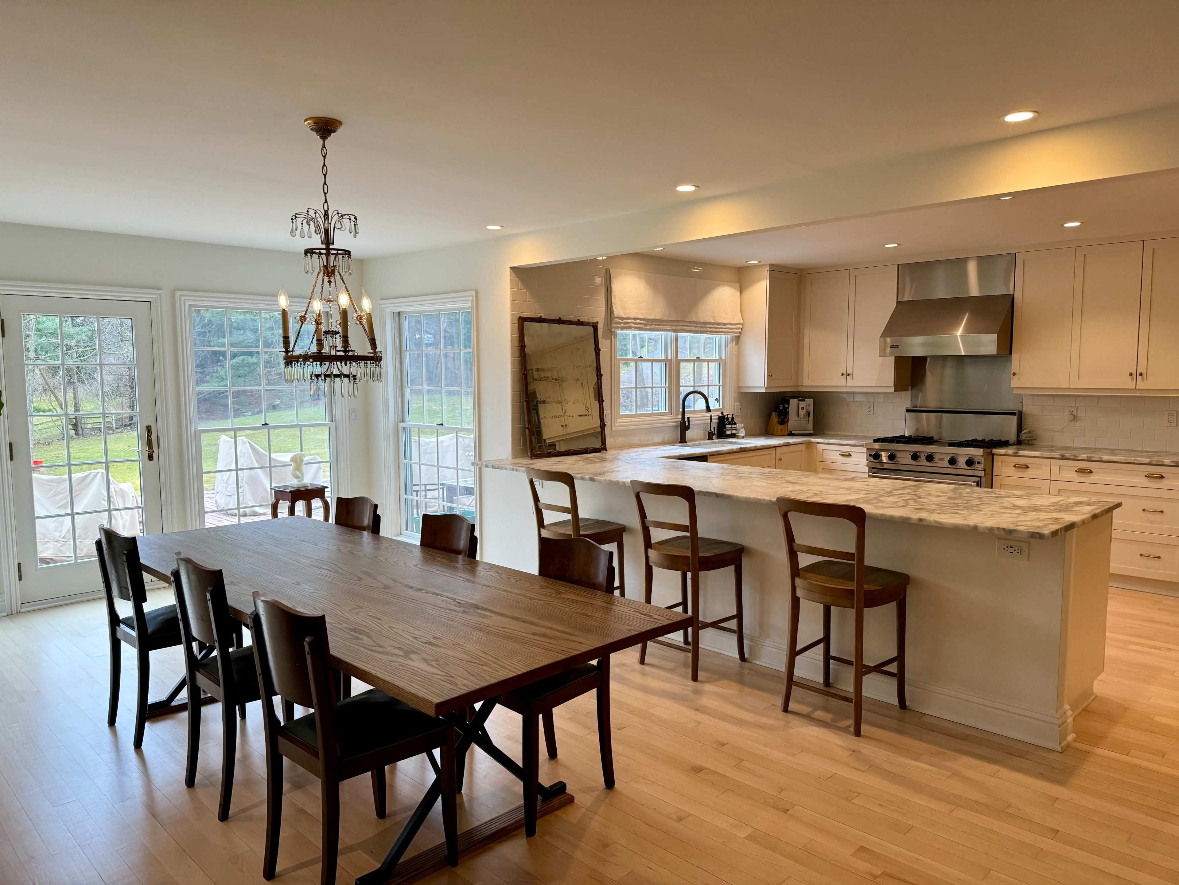 A spacious kitchen and dining area features a large wooden table with several chairs, an island with bar stools, and large windows providing a view of the outdoors.