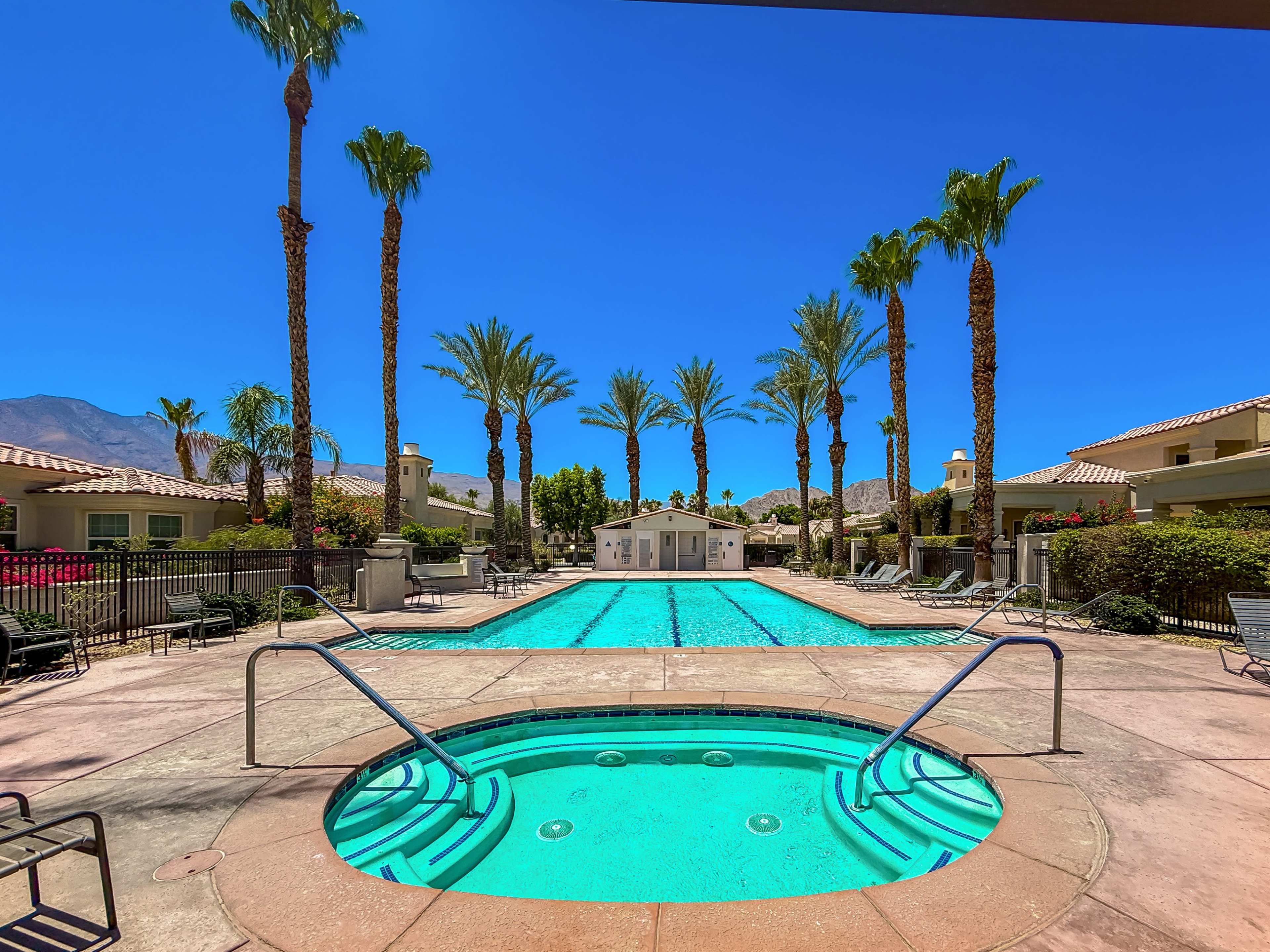 The image shows a swimming pool area surrounded by palm trees and residential buildings under a clear blue sky.