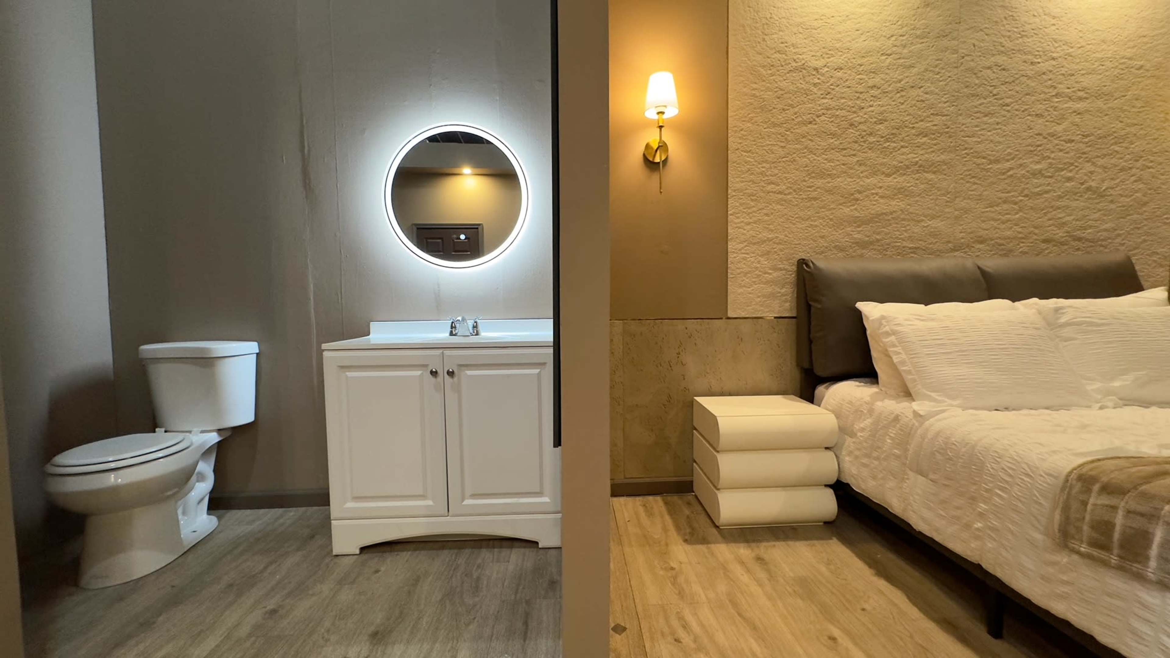 A split view of a bathroom on the left with a toilet and sink, and a bedroom on the right featuring a bed and nightstand.
