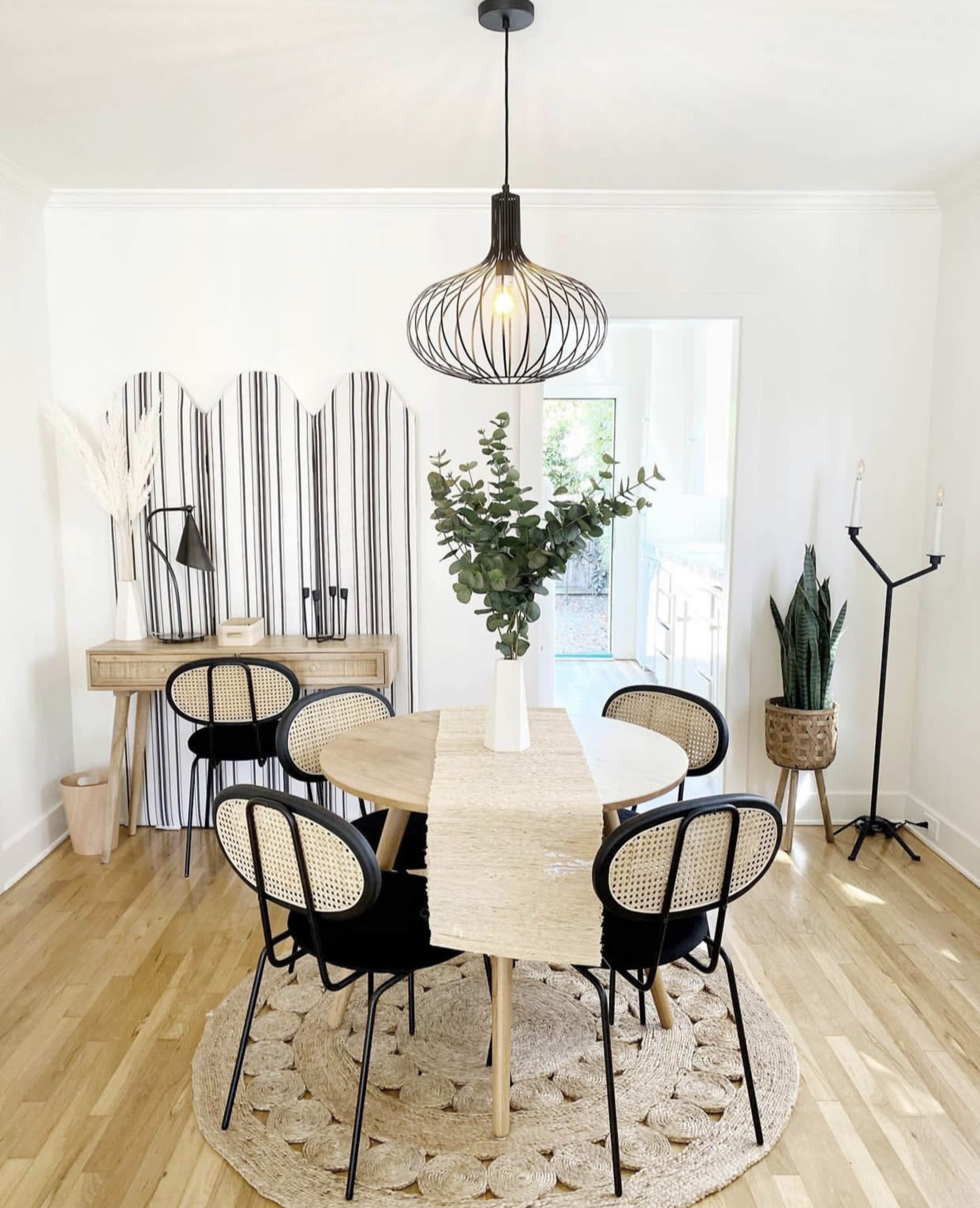 A modern dining area features a round table surrounded by six black and rattan chairs, with a decorative vase of greenery on the table and a patterned area rug beneath it.