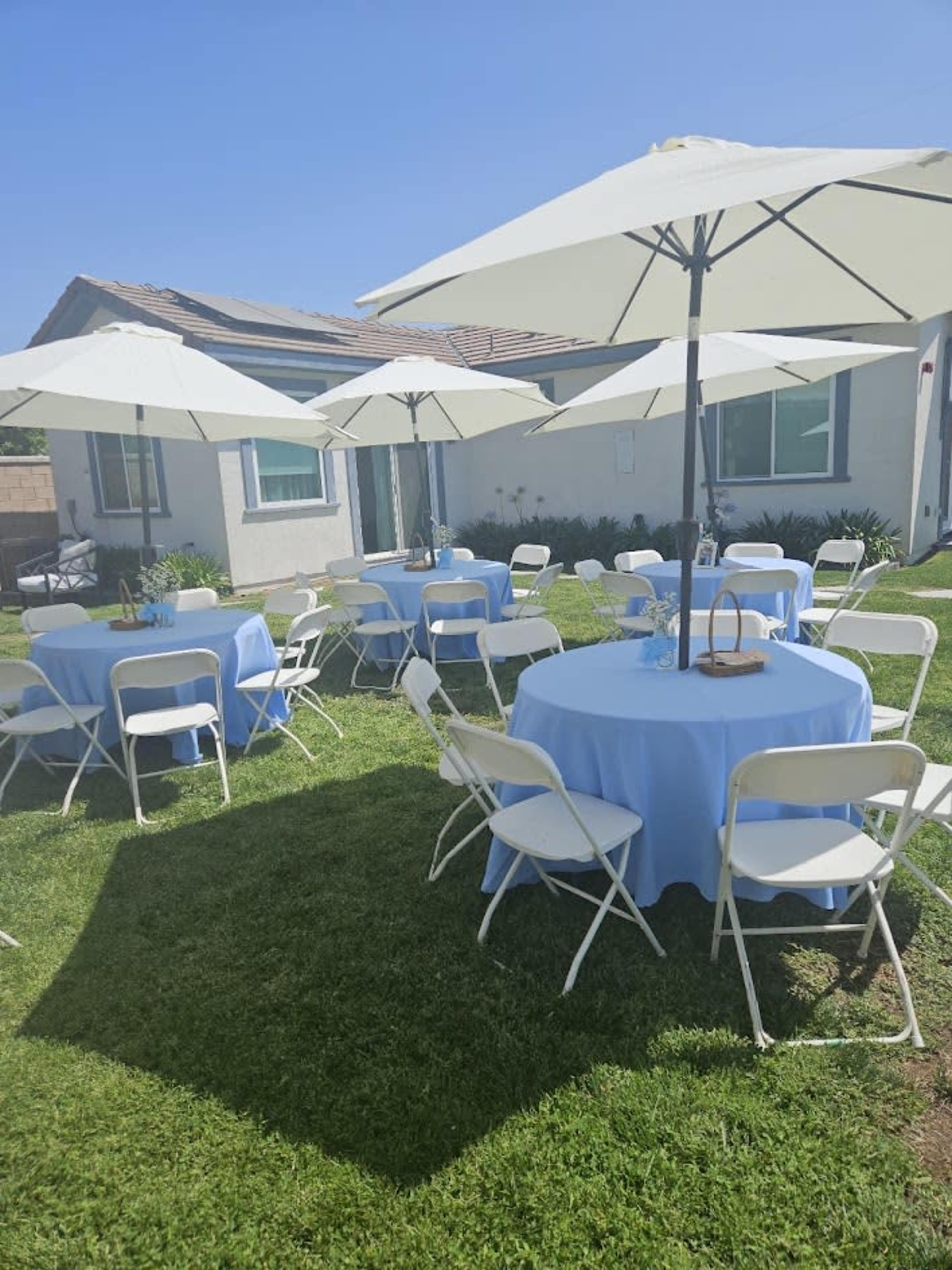 The image shows a neatly arranged outdoor event space with several round tables covered in light blue tablecloths and white umbrellas providing shade on a grassy area.