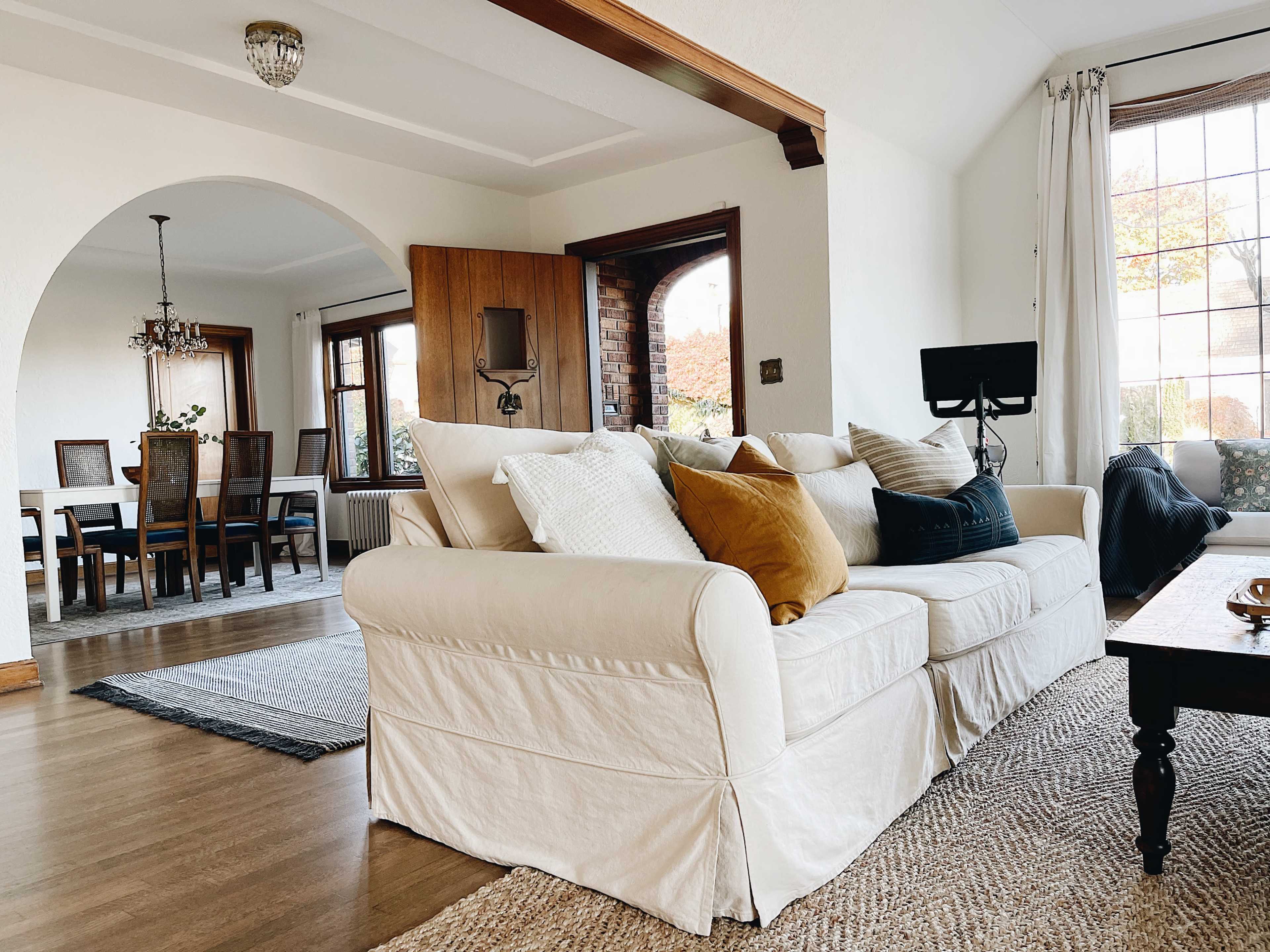 A light-colored sofa with various cushions is positioned in a bright living room that connects to a dining area featuring a wooden table and chairs.