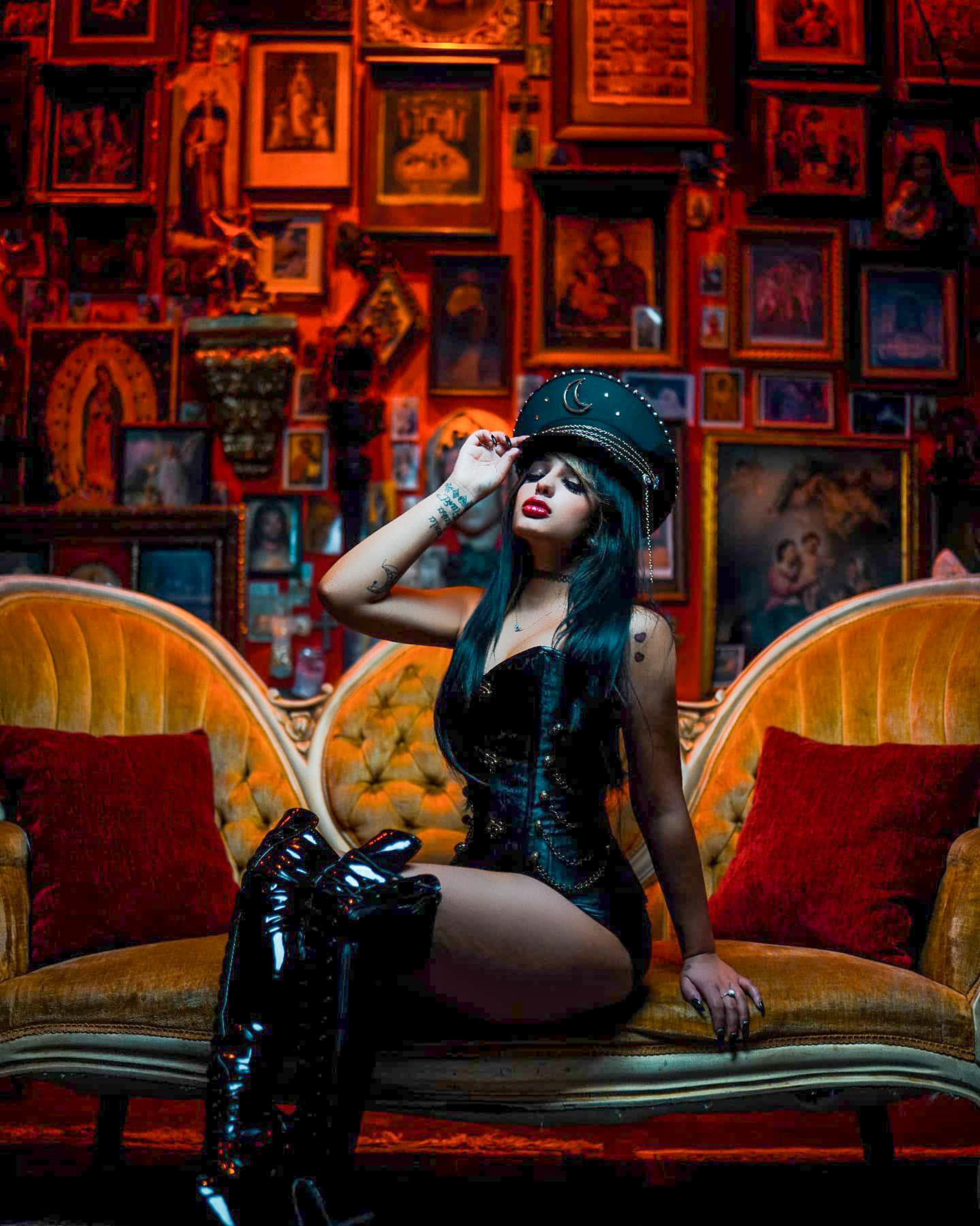 A woman in a black corset and knee-high boots sits on a velvet sofa in front of a wall adorned with numerous framed pictures.