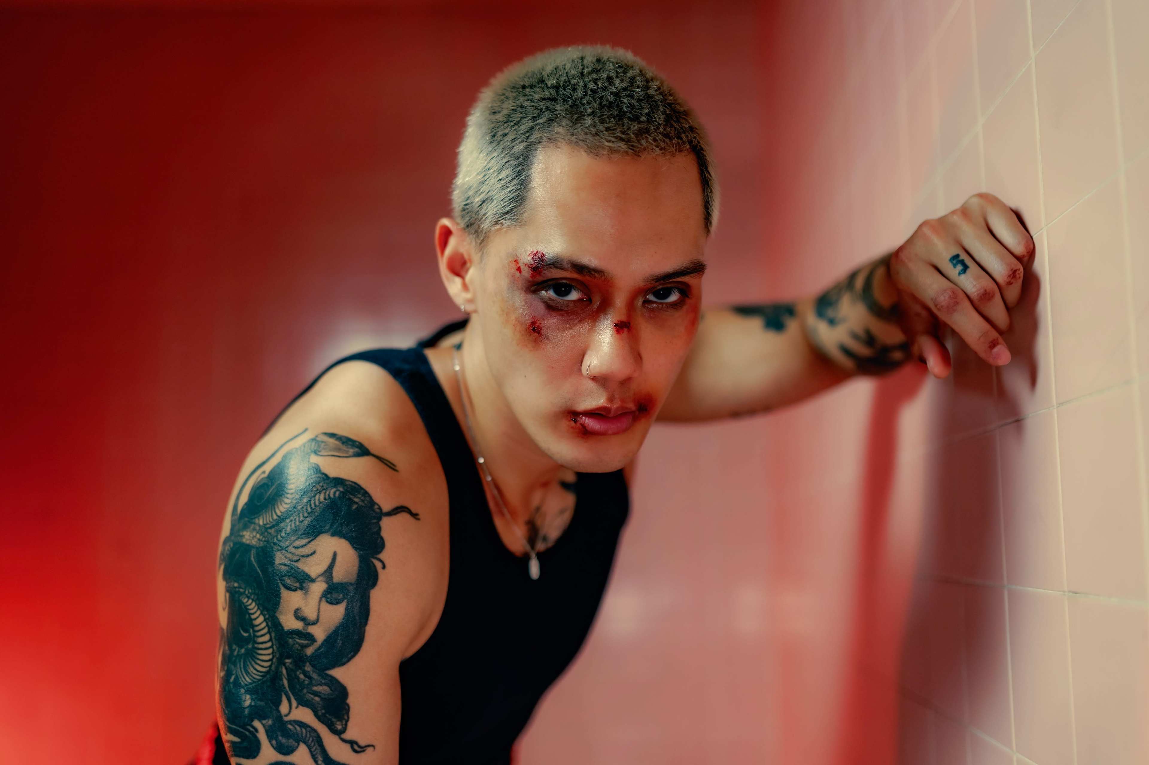 A person with short, light-colored hair and visible tattoos leans against a pink wall, displaying signs of injury on their face.