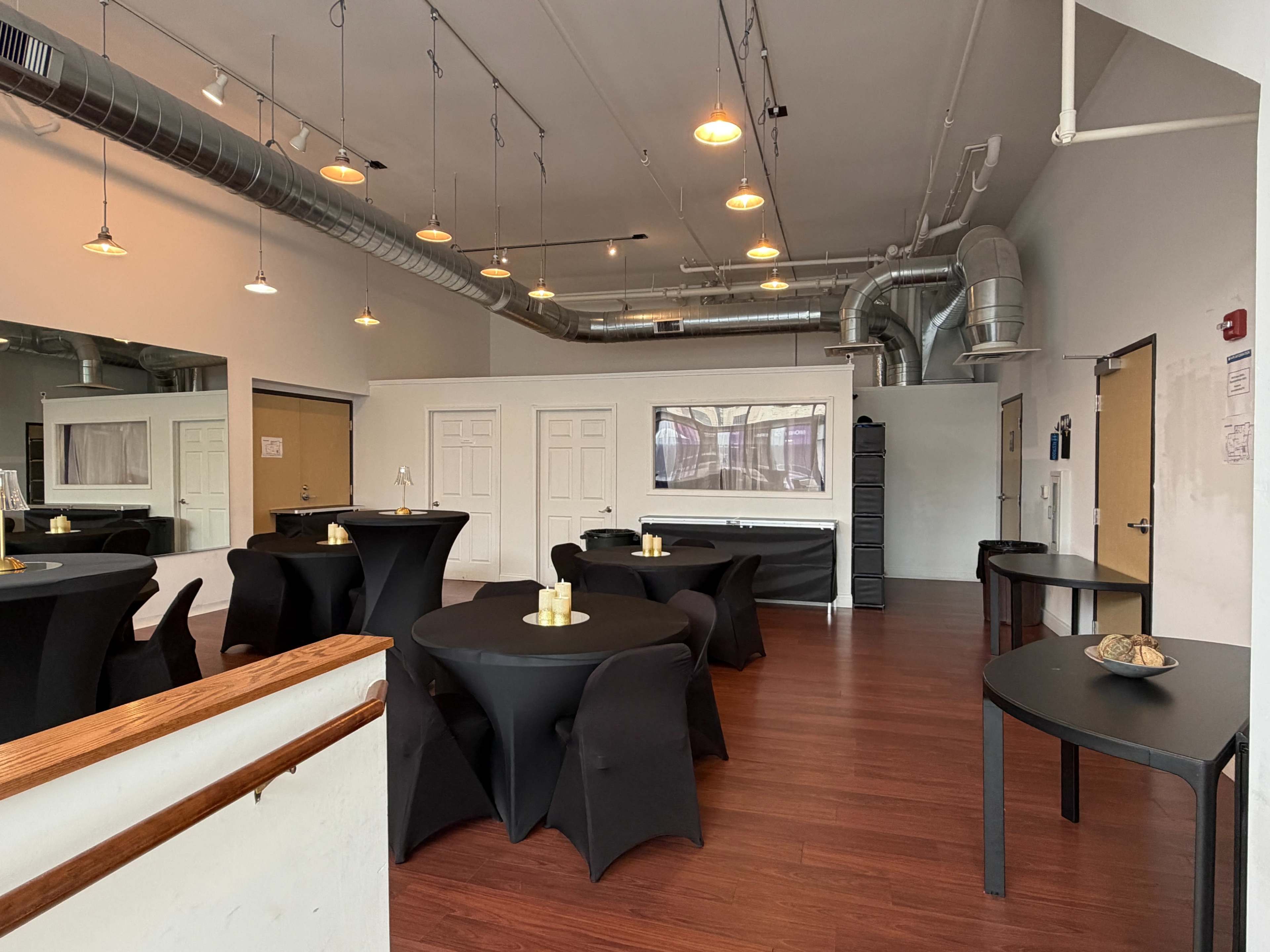 The image shows a spacious event room with several black, round tables, some topped with small candles, and a bar area at the back with white walls and exposed ductwork above.