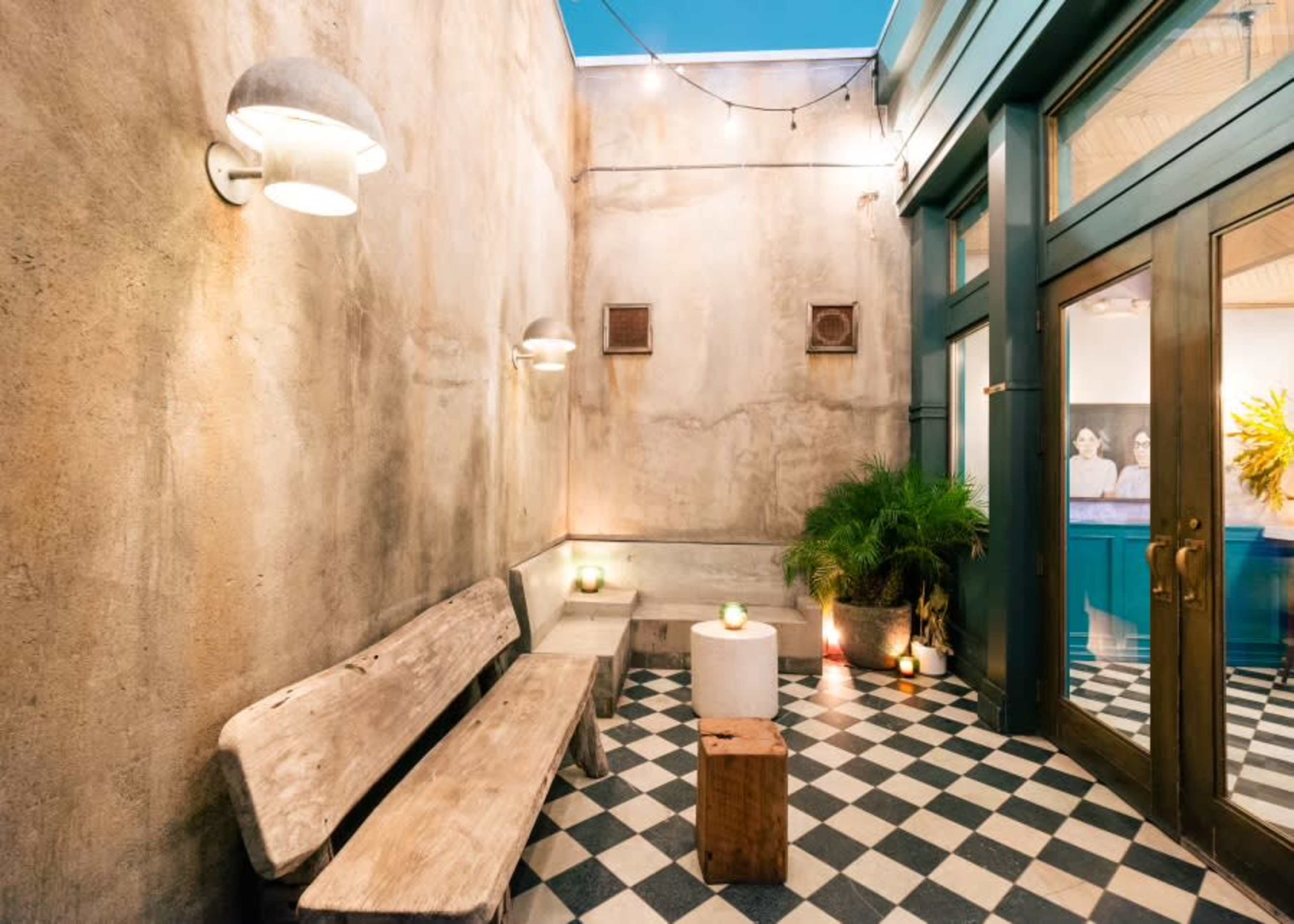 The image shows an outdoor corner with a textured wall, checkered tile flooring, wooden benches, and soft lighting.