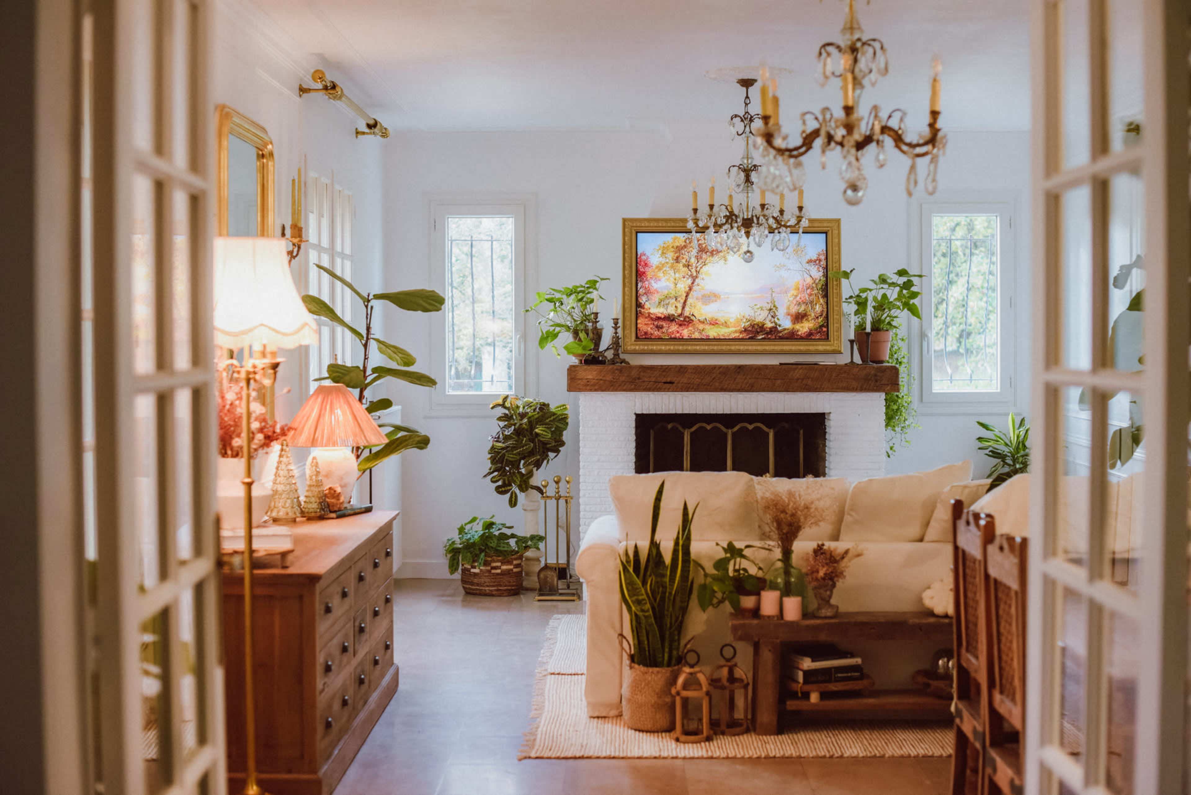 The image shows a living room with a white sofa, a wooden coffee table, a fireplace adorned with plants, and a chandelier hanging from the ceiling.