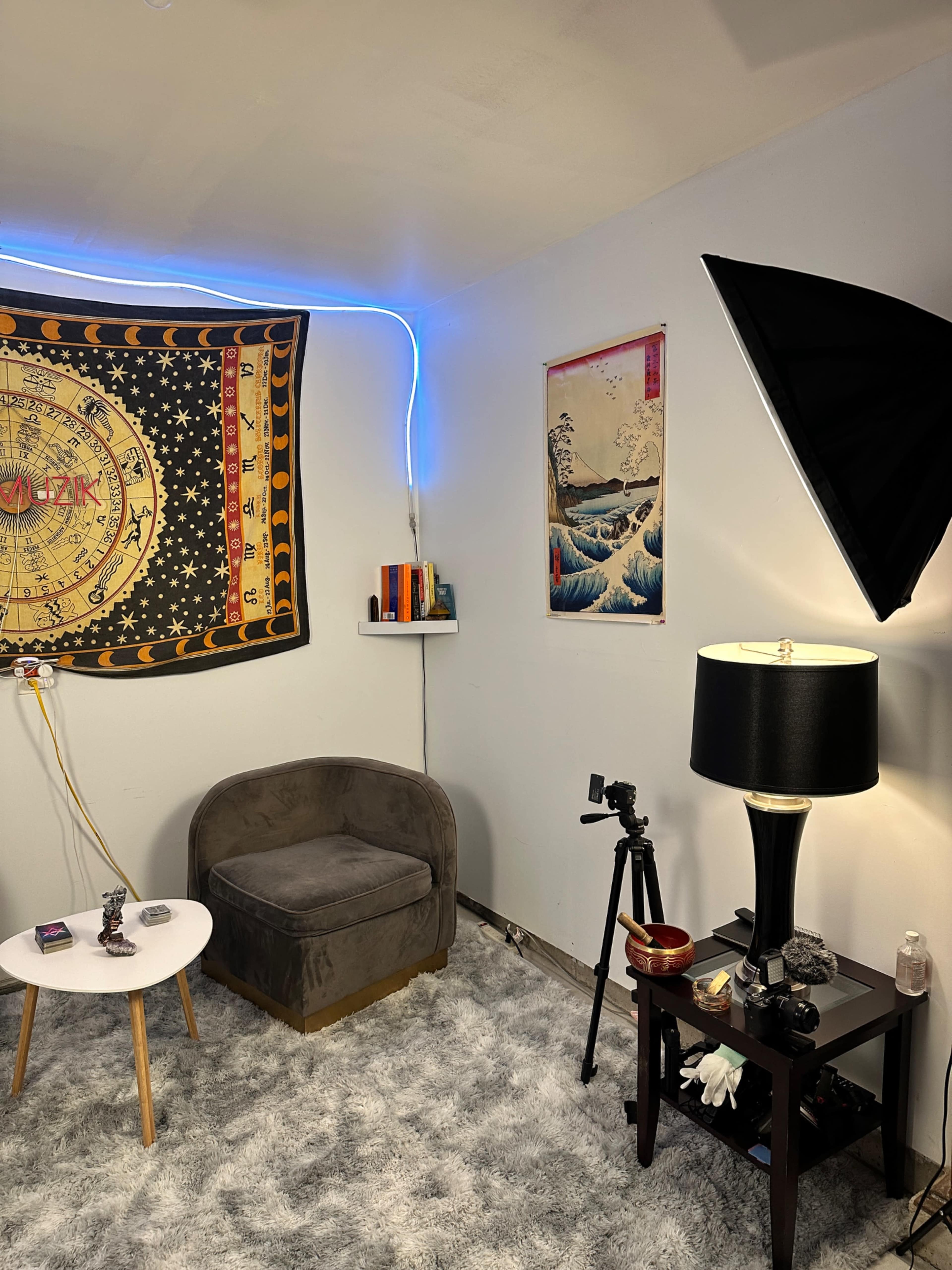 The image shows a cozy room with a leather chair, a small round table, a decorative wall tapestry, a mounted light source, and a tripod with a camera on one side.