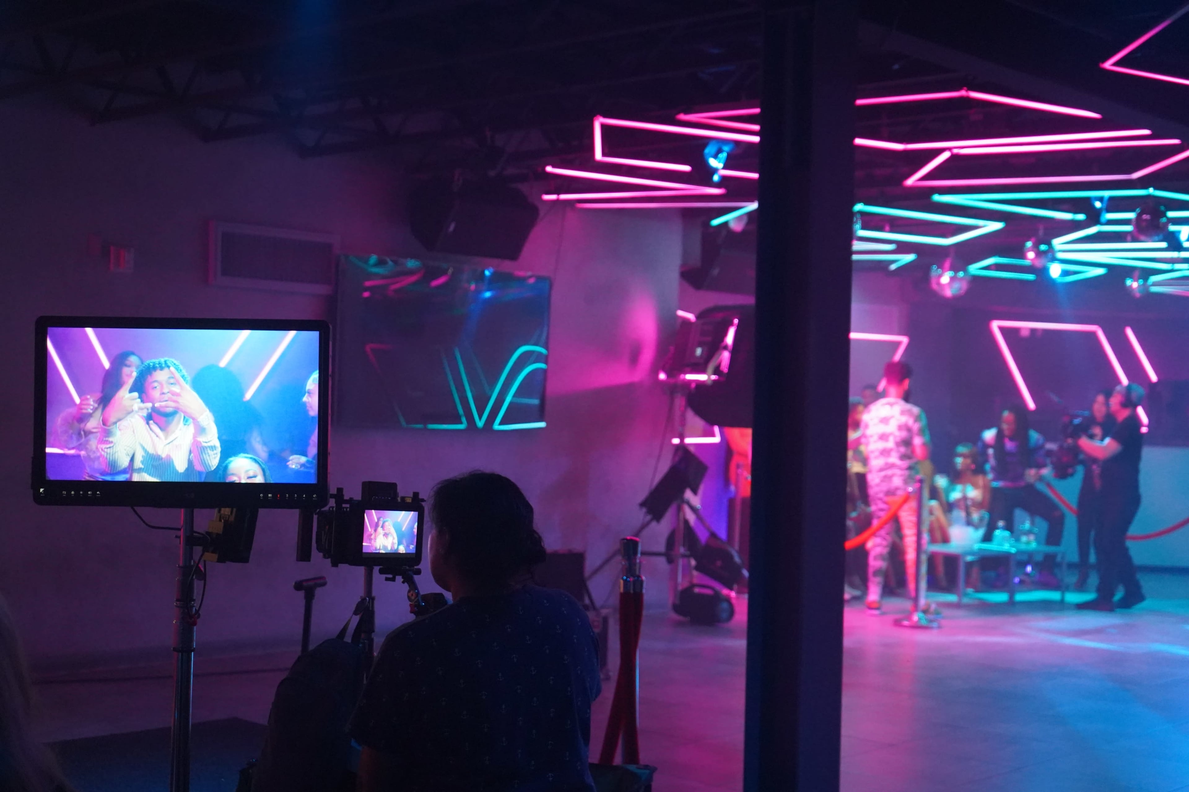 A film crew captures a colorful scene in a nightclub setting with neon lighting and multiple cameras positioned on tripods.