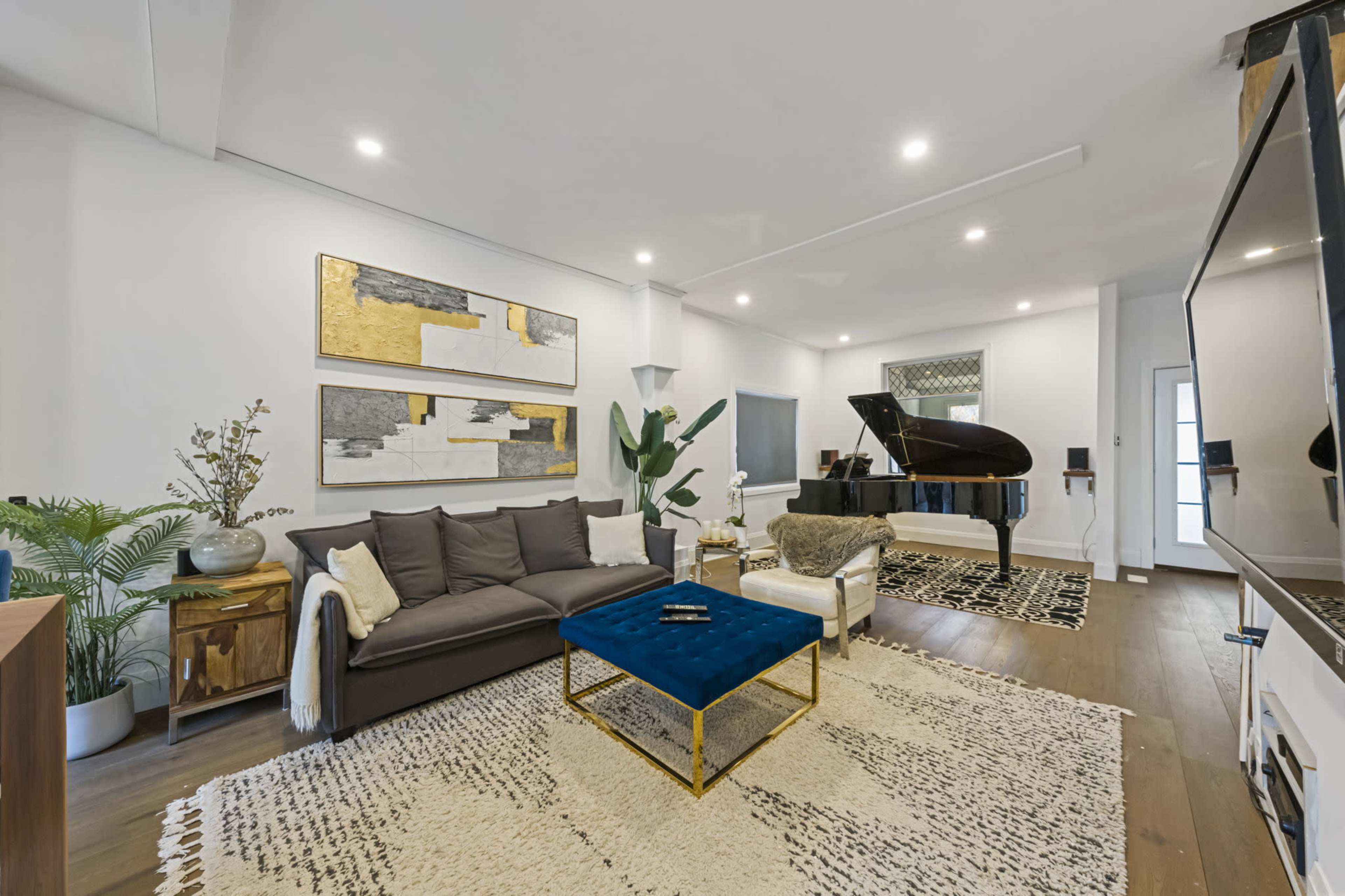A modern living room features a black piano, a blue coffee table, a large rug, and abstract wall art against white walls.
