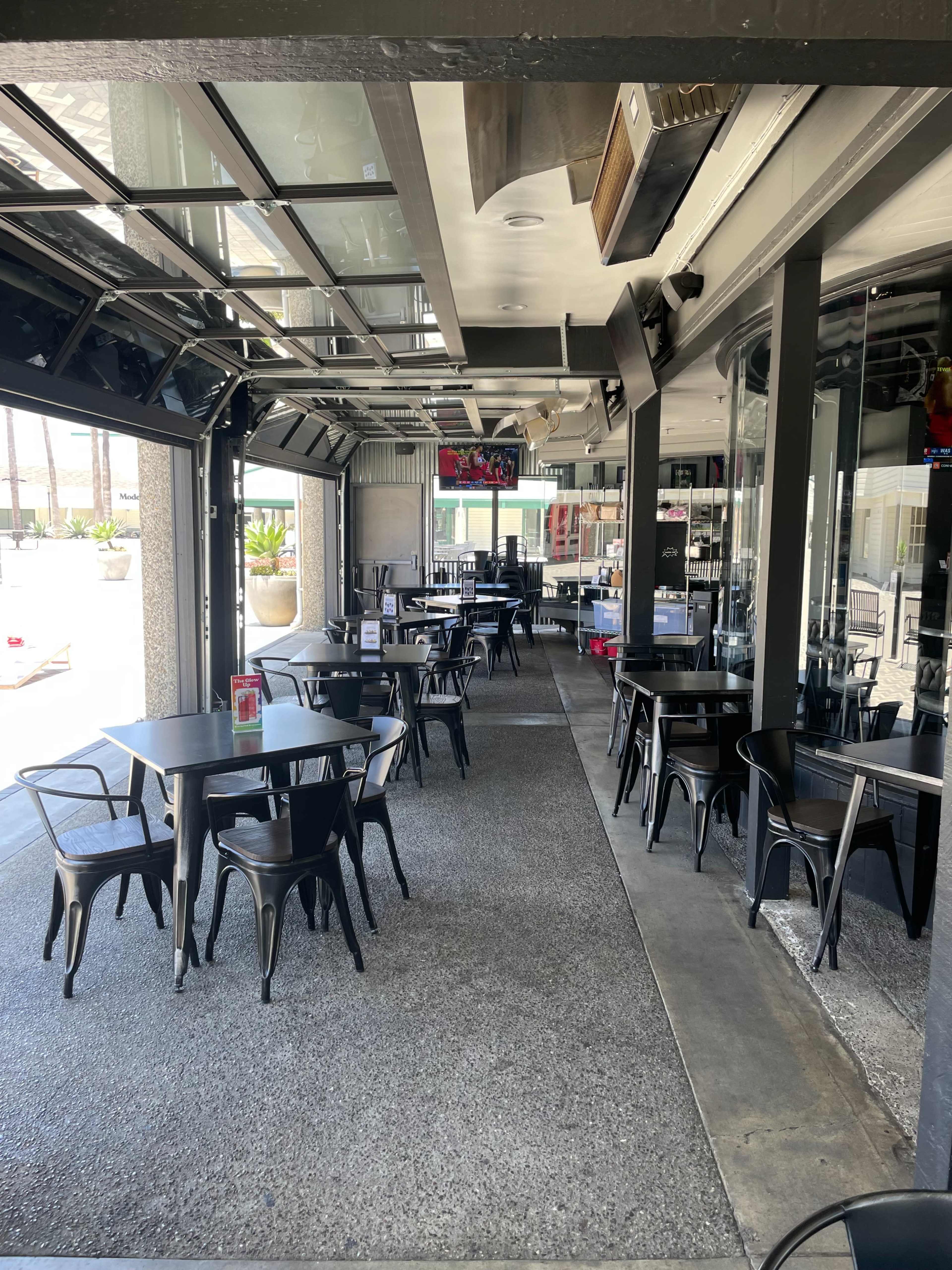 The image shows an empty restaurant interior with black tables and chairs arranged along a walkway, featuring large glass doors that open to the outside.