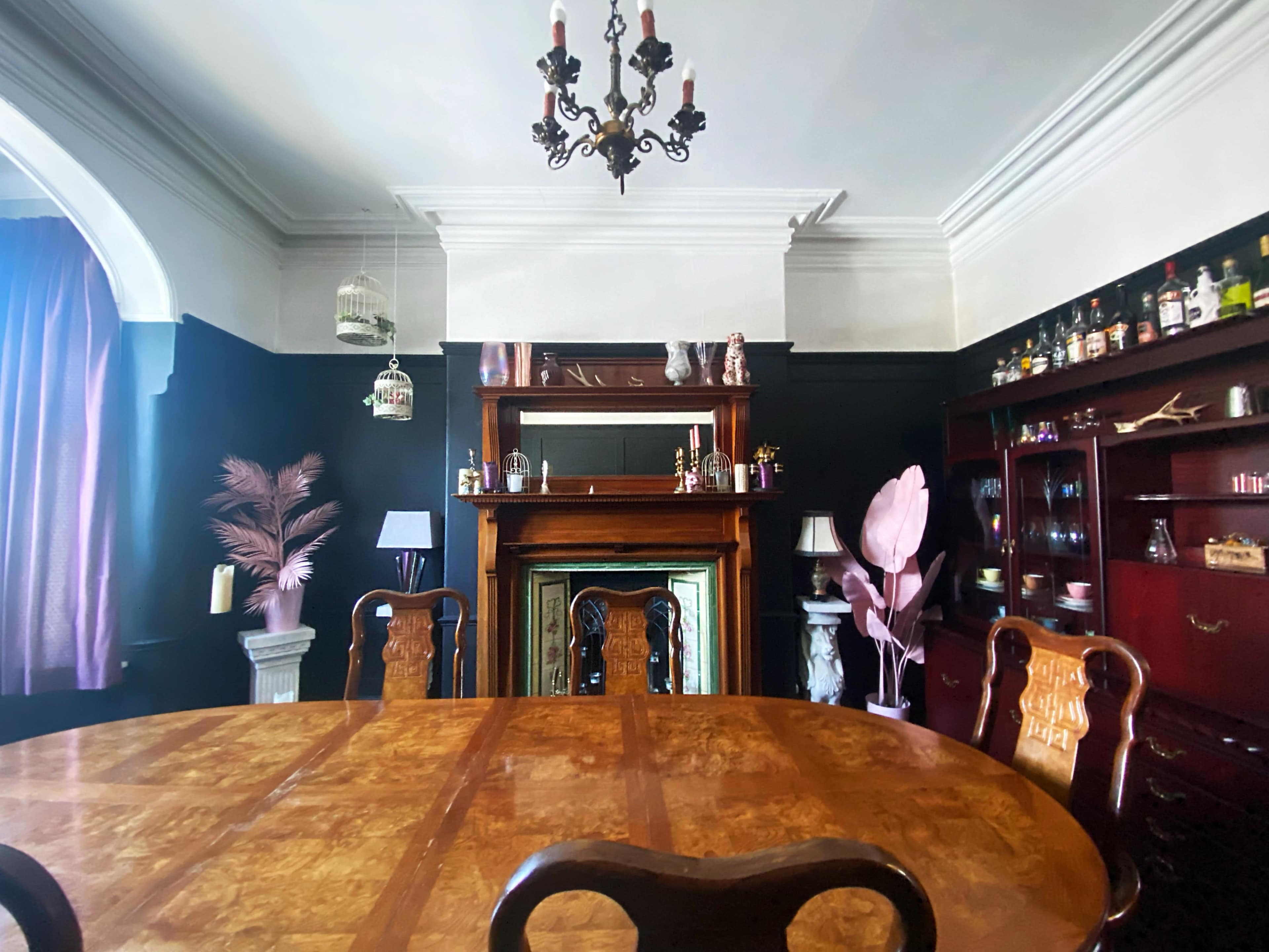 A wooden dining table is positioned in a room with dark walls, featuring a fireplace, a mirror, and various decorative items.