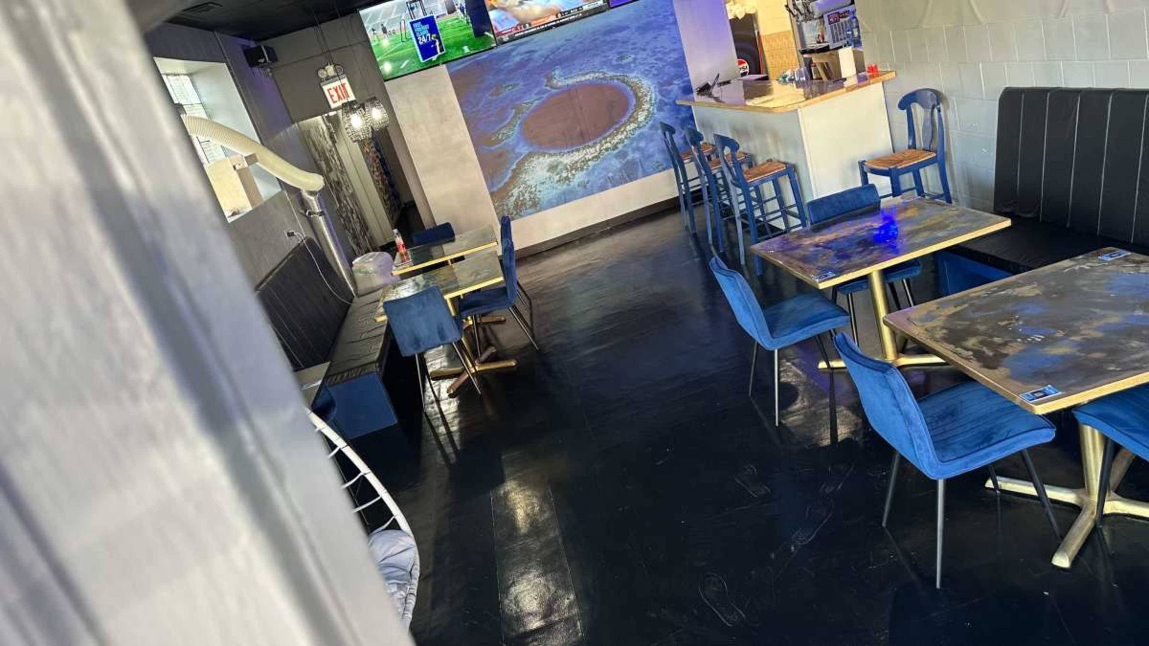 The image shows a modern restaurant interior with blue chairs and tables, a bar area, and a large screen displaying a video.