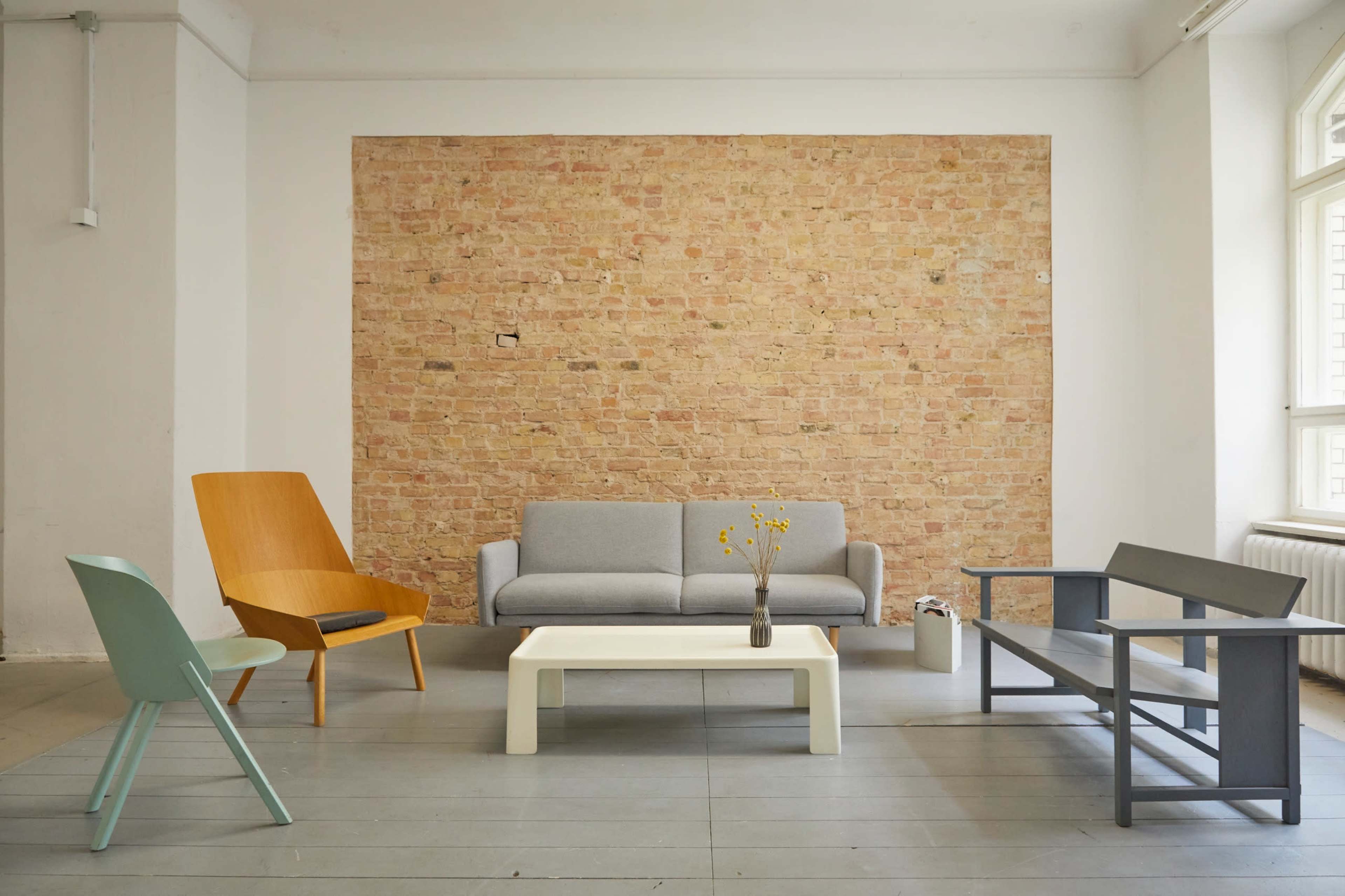 A minimalist living room with a gray sofa, an orange accent chair, a white coffee table, and a gray bench against a brick wall.