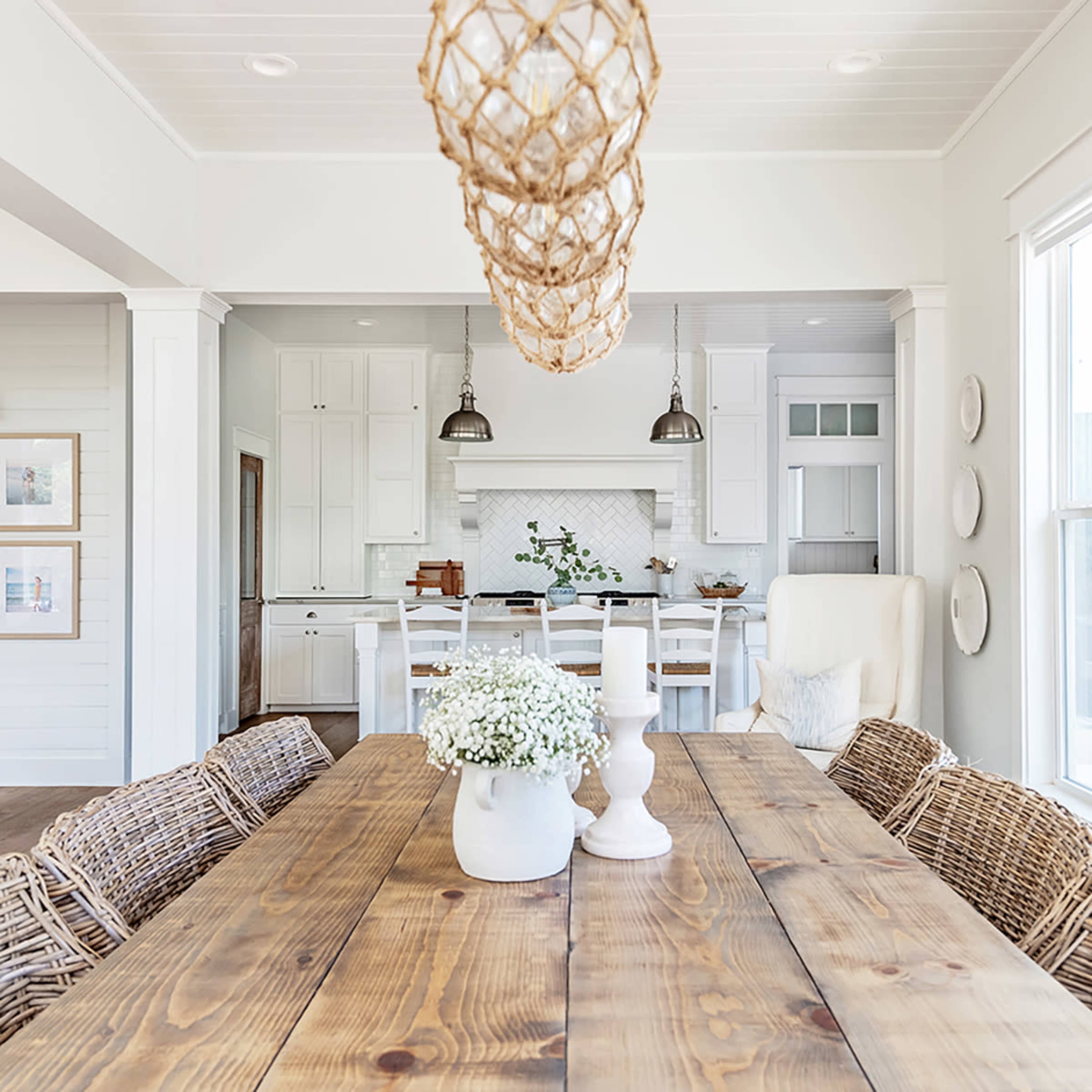 A wooden dining table with a vase of flowers is set under a pendant light in a bright, open kitchen space with white cabinetry and a light tile backsplash.