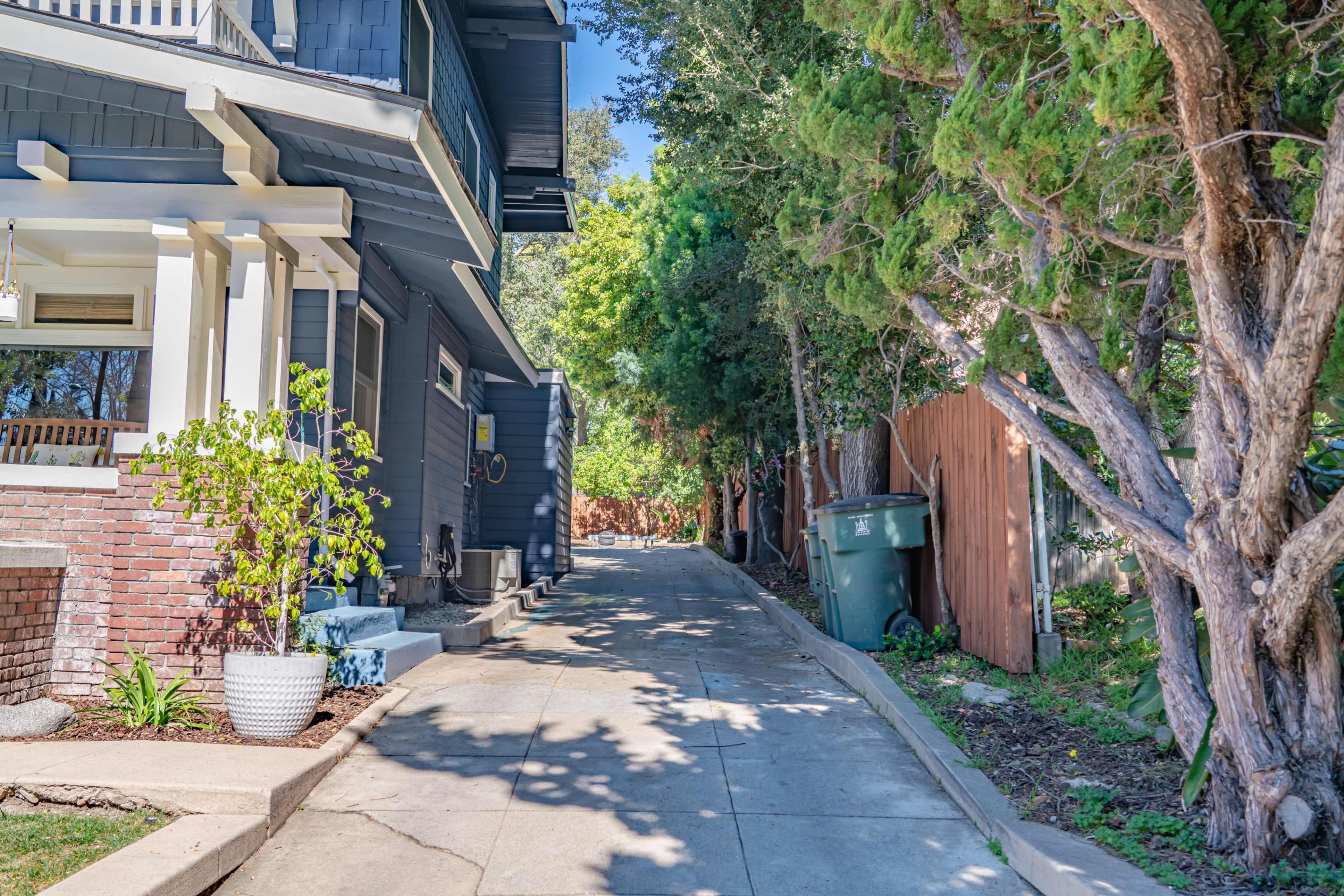 A narrow concrete pathway runs between a house and a fence, lined with trees and a green trash bin.