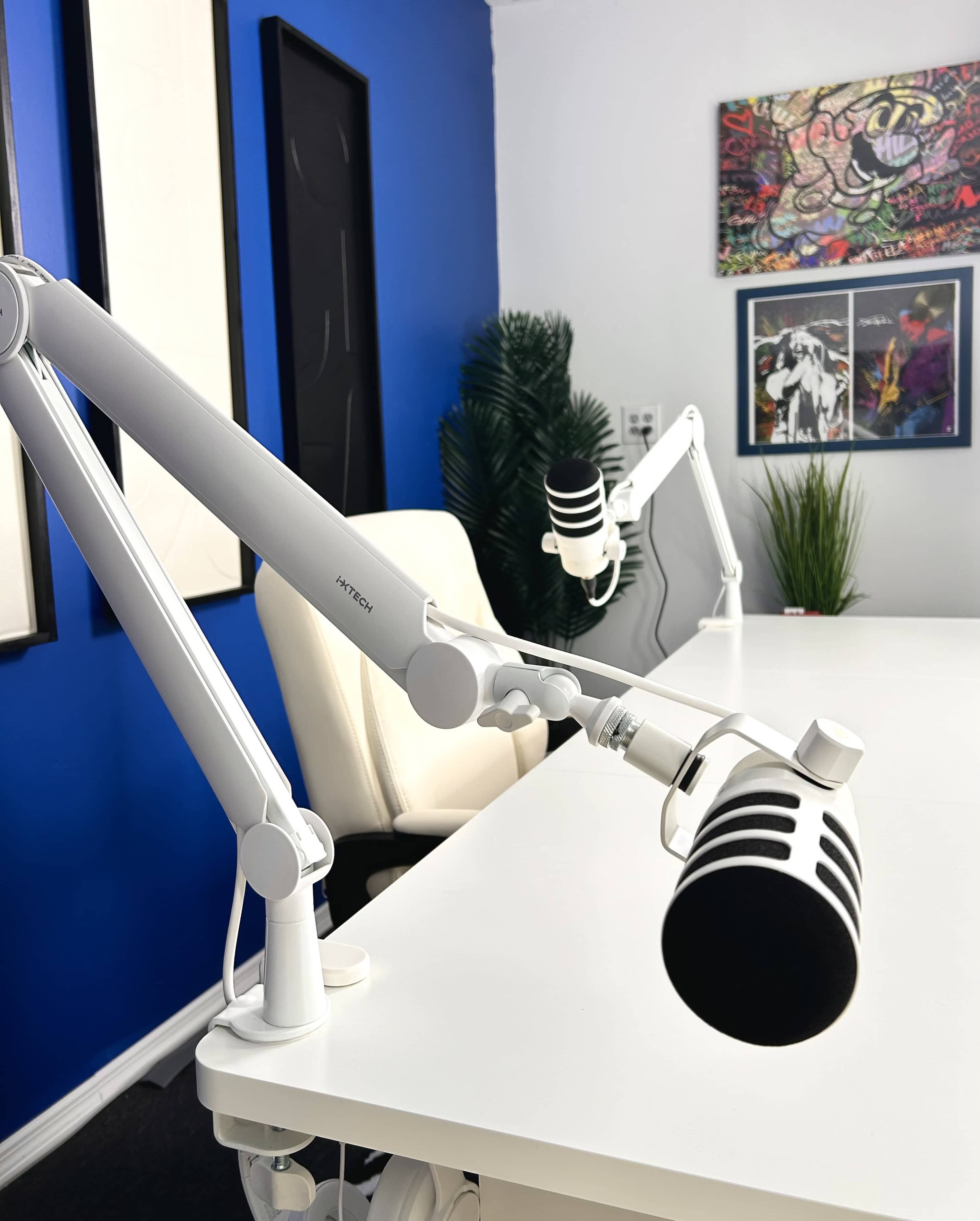The image shows a well-lit podcast studio featuring white adjustable microphone arms, a sleek white table, and colorful artwork on the walls.