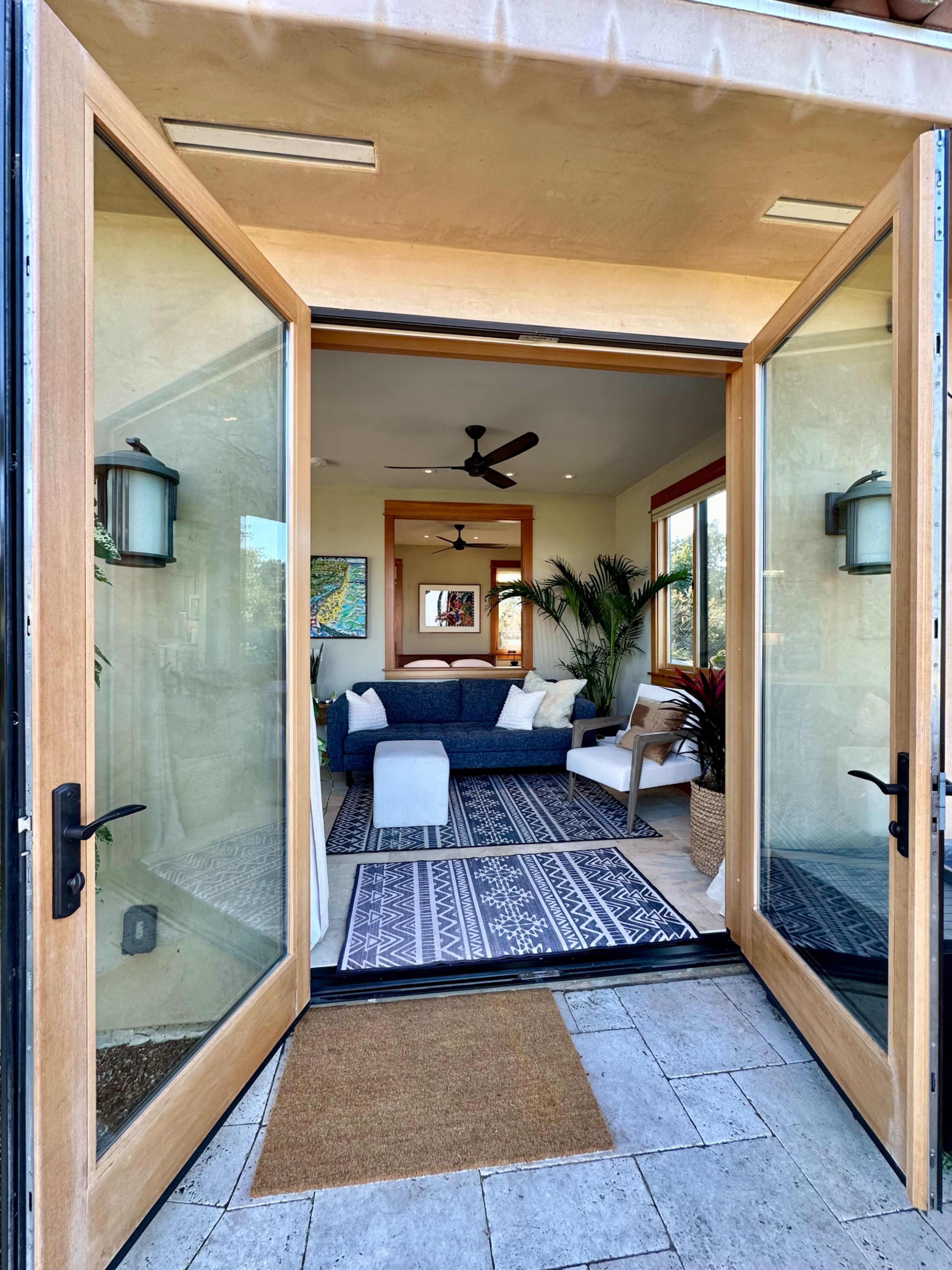 A pair of large glass doors opens to a bright, modern living room with a rug, a sofa, and tropical plants.