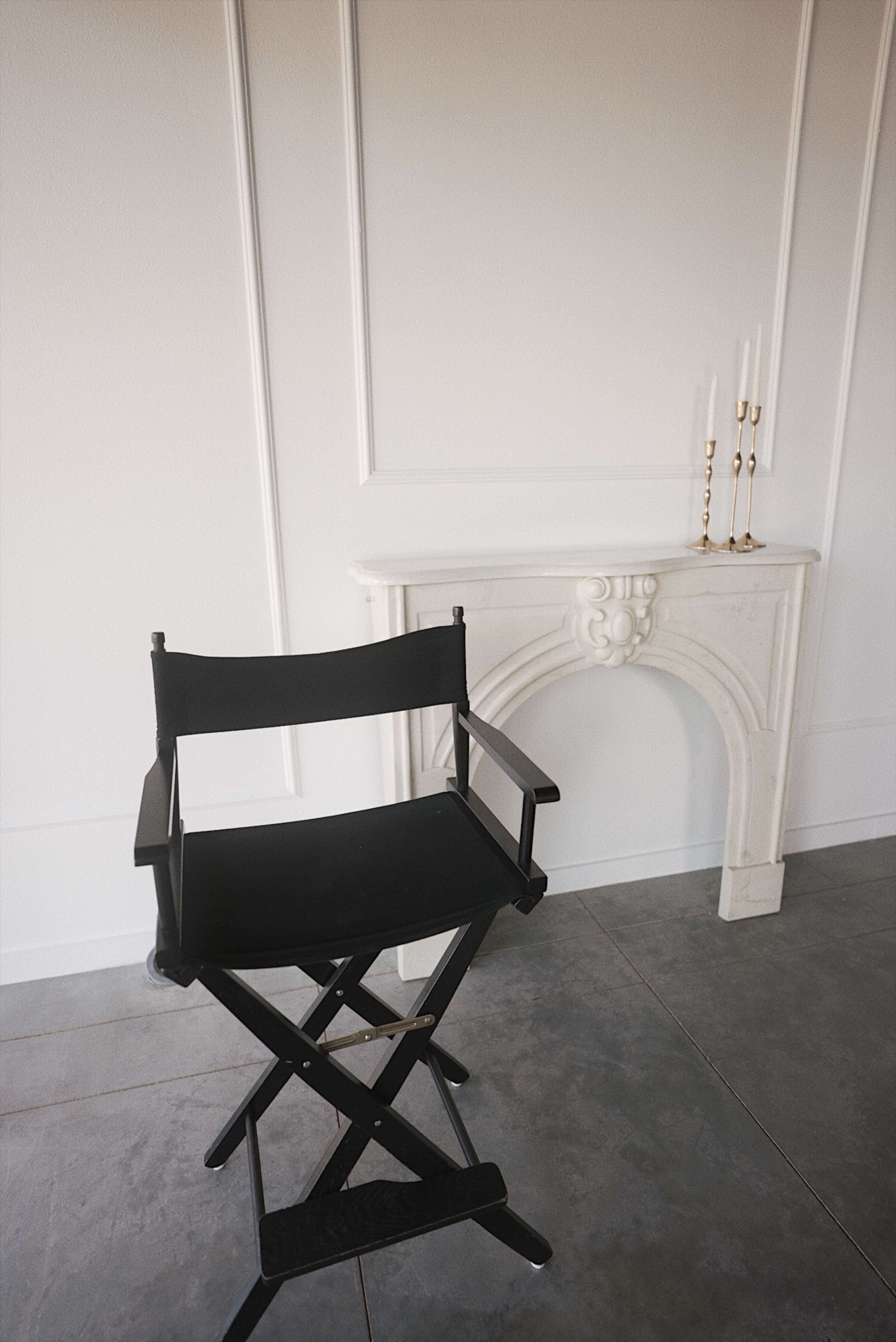 A black director's chair stands next to a white marble fireplace adorned with three slender candlesticks.