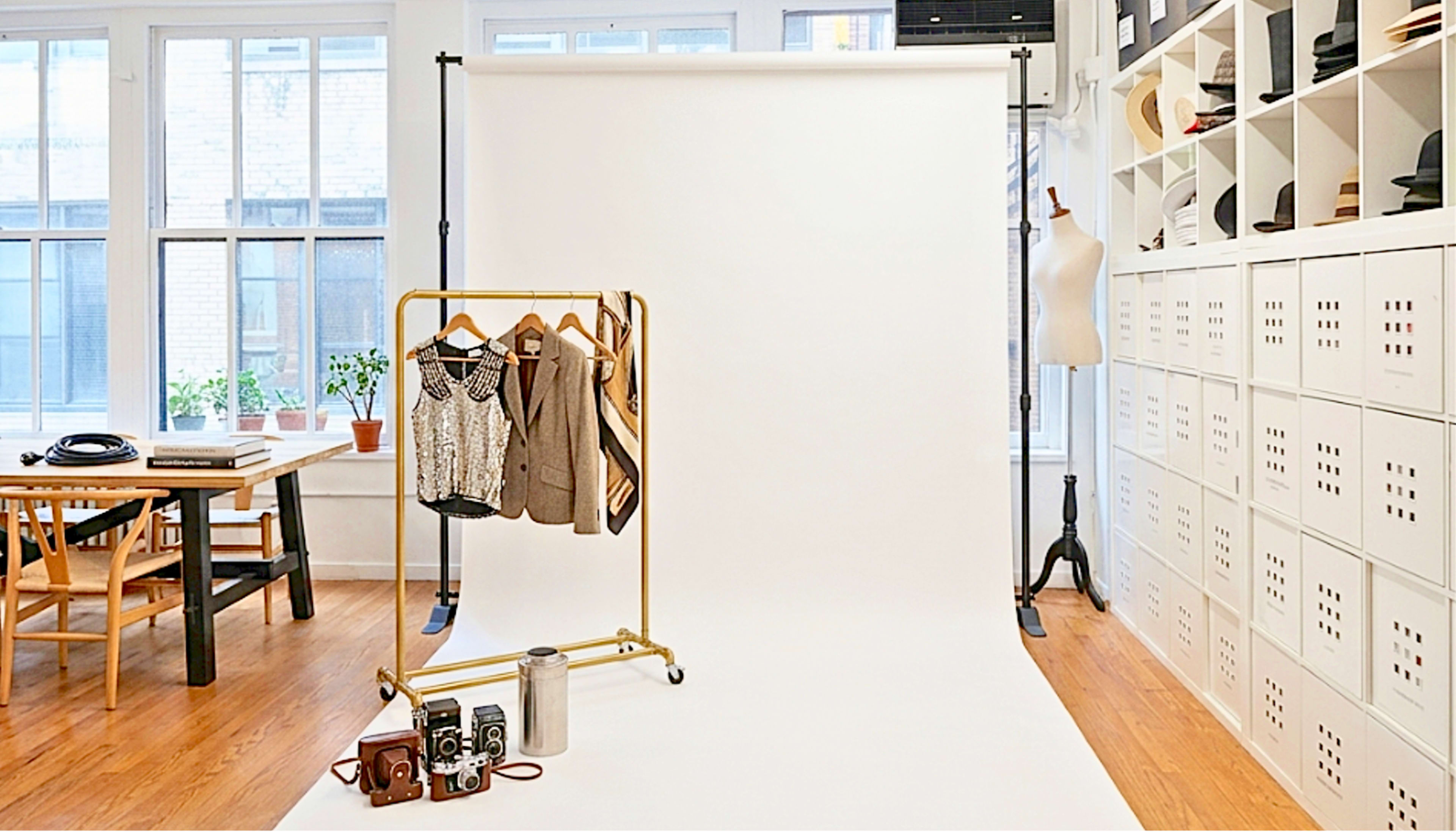 The image shows a photography studio with a white backdrop, a clothing rack displaying outfits, a vintage camera, and a thermos on the floor, alongside a table and storage units.