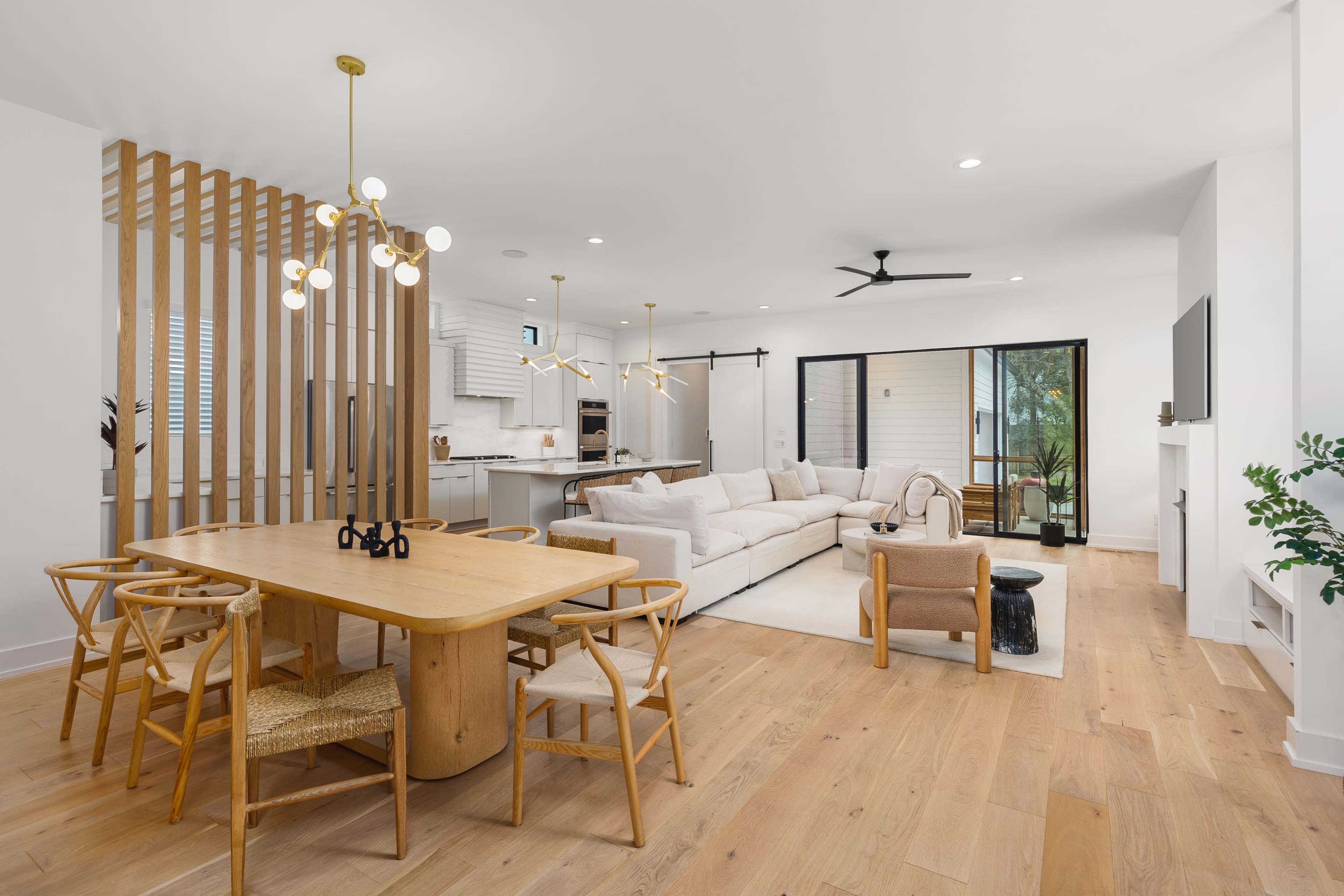 The image shows a modern living space featuring a light-colored sofa, a wooden dining table with chairs, and large windows that open to a view of greenery.