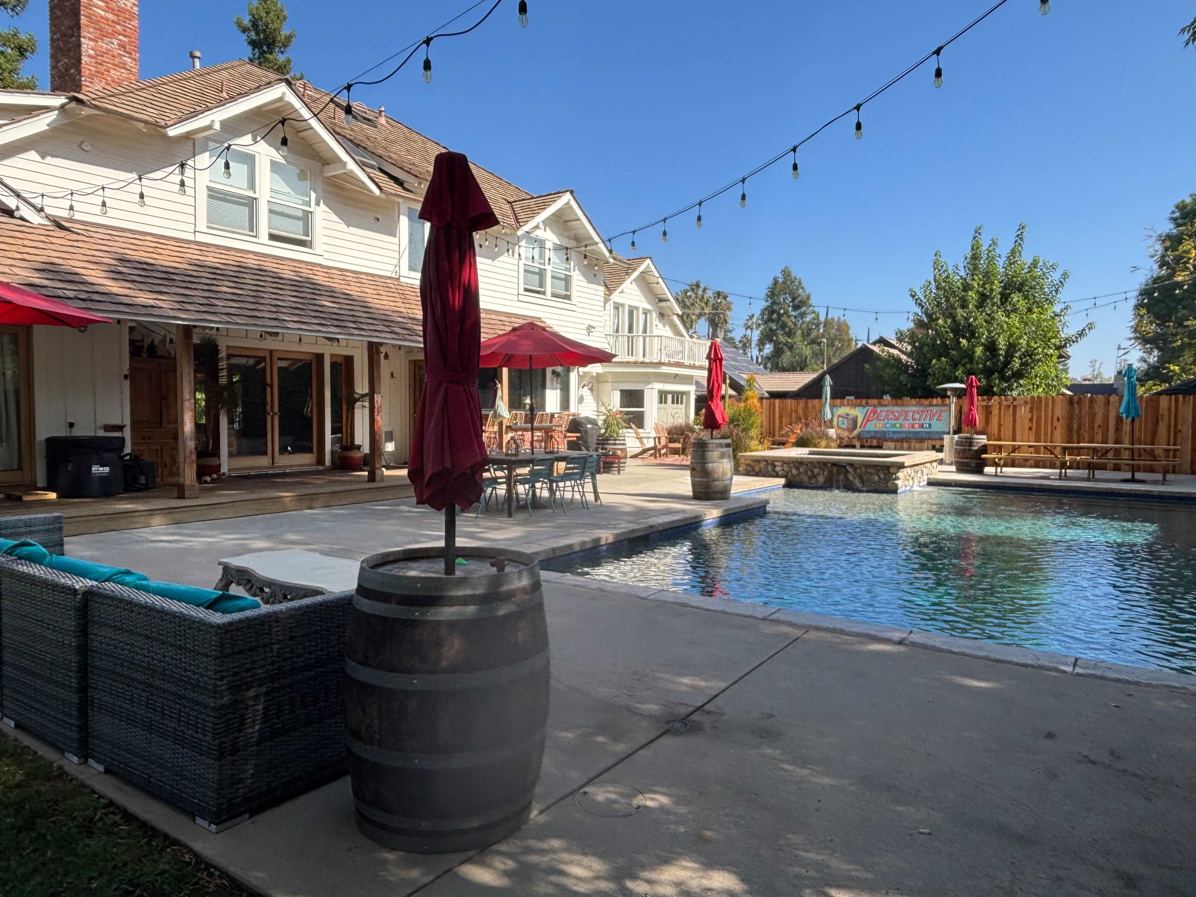 A backyard scene features a pool surrounded by patio seating, a wooden deck, and umbrellas, with a house in the background.