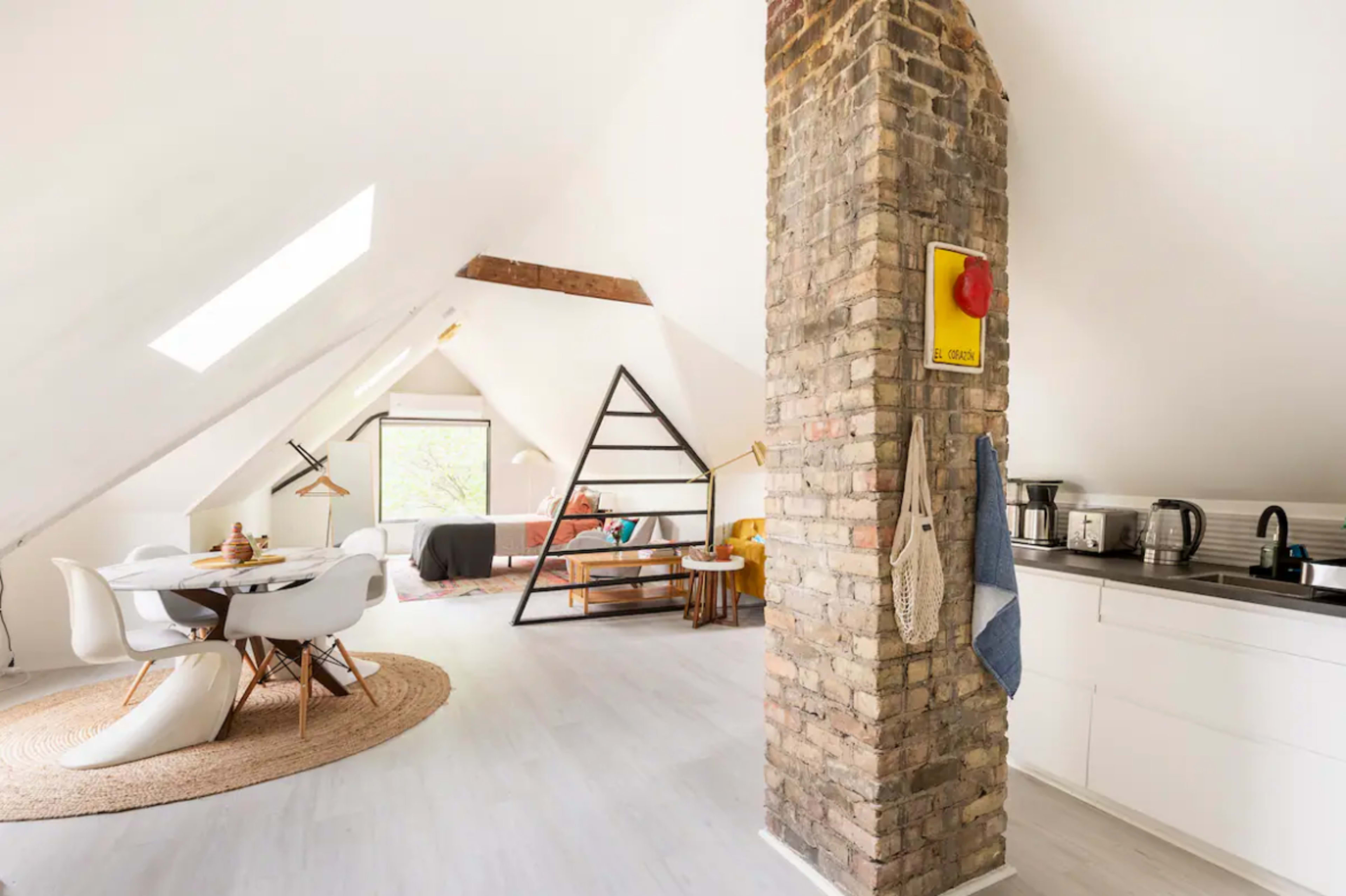 A modern attic apartment featuring a kitchen area, dining space with white chairs, and a cozy living area with a distinct brick column and skylights.