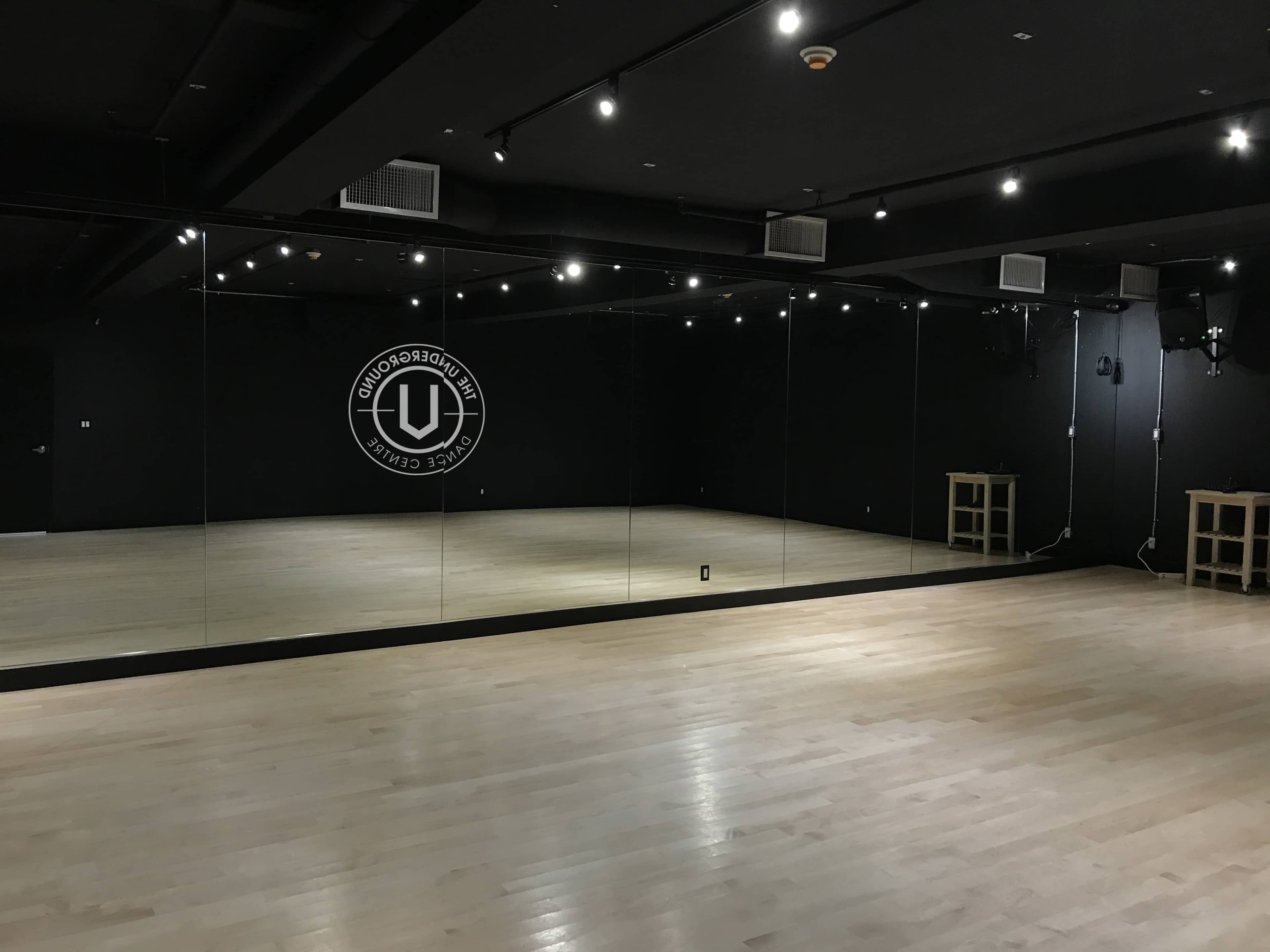 The image shows a spacious dance studio with mirrored walls and hardwood flooring, illuminated by overhead lights.