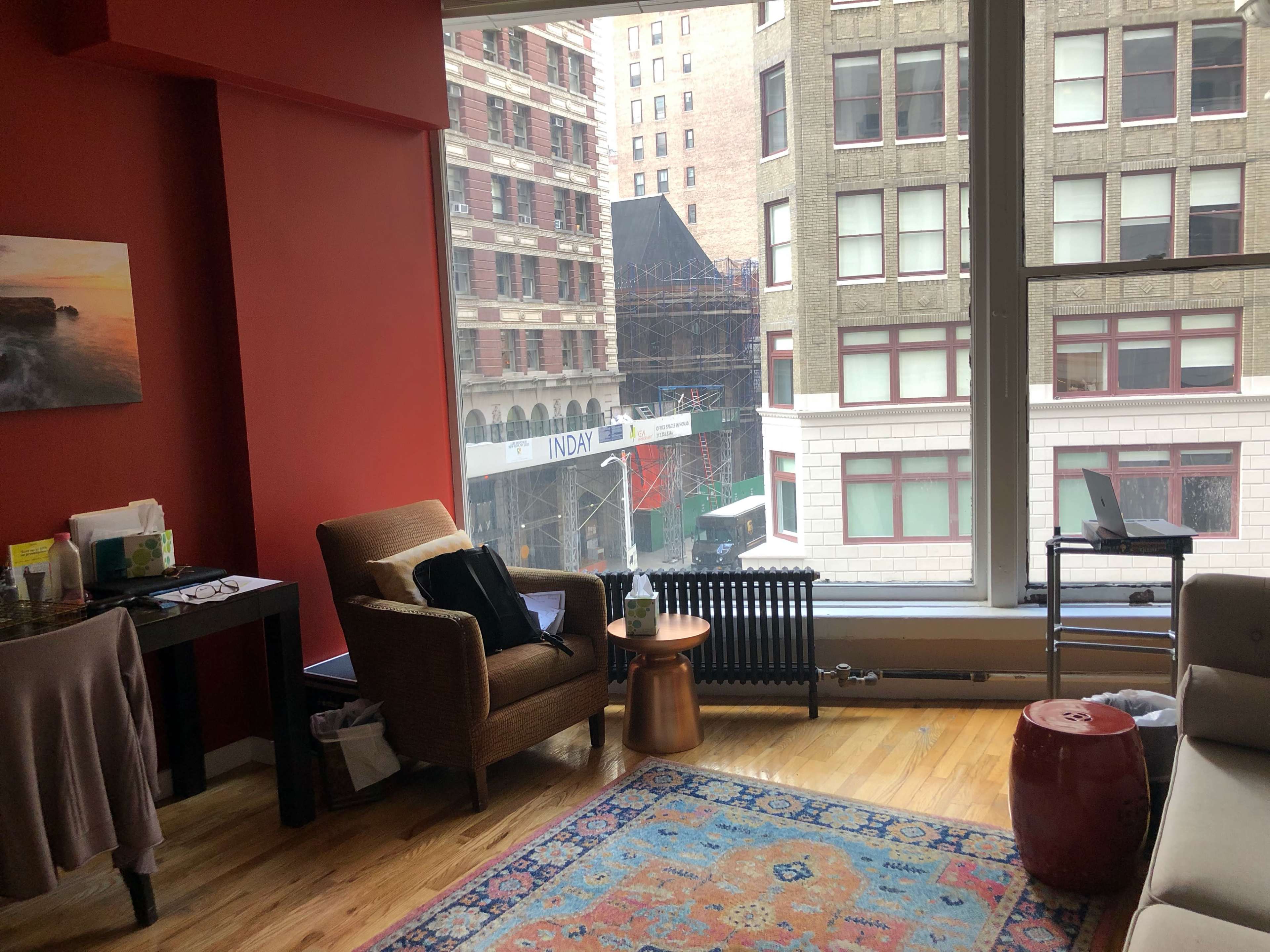 A cozy living room features a chair and a table beside a large window overlooking an urban street with buildings and construction activity outside.
