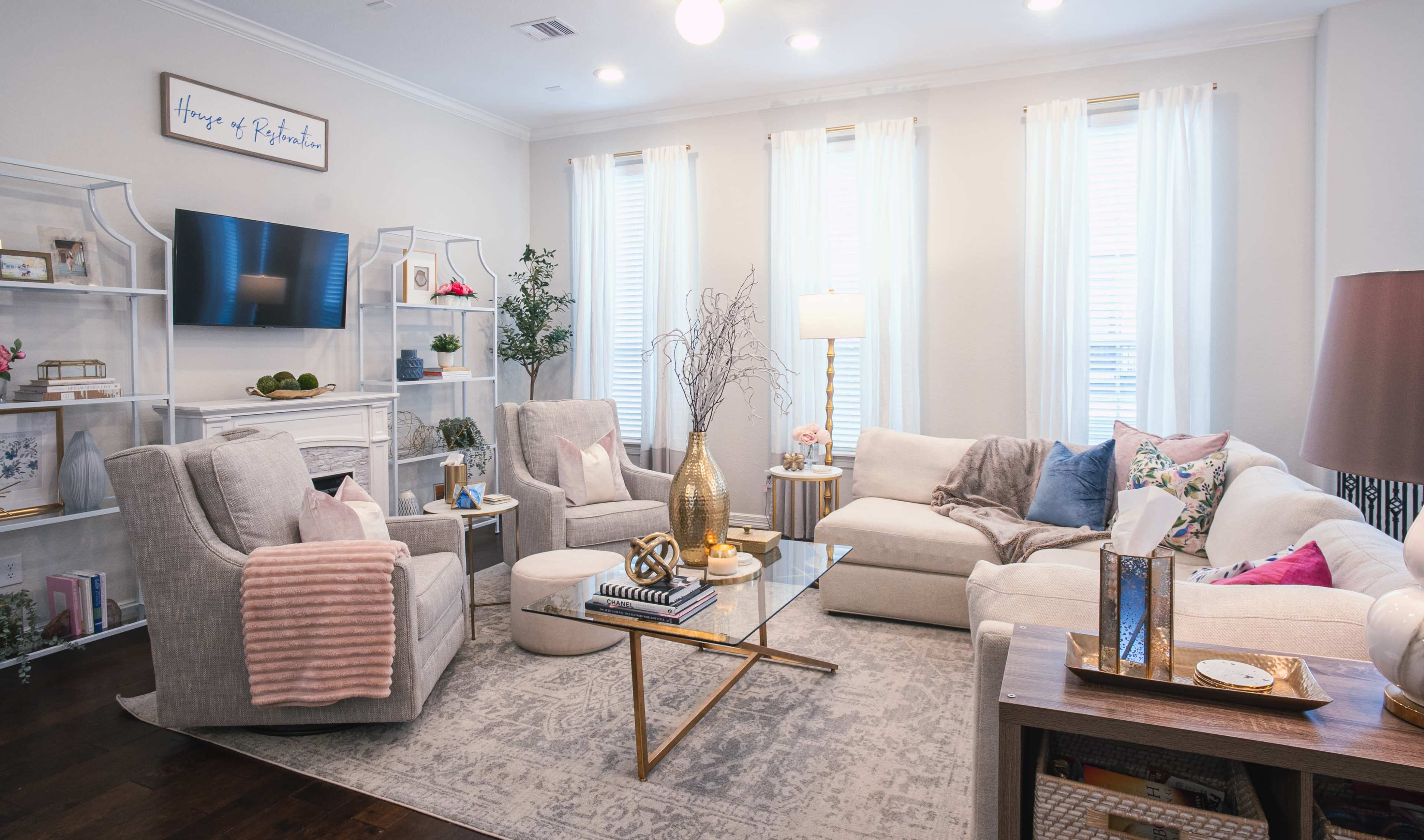 A modern living room features a cream-colored sectional sofa, two light gray armchairs, a glass coffee table, and a white bookshelf adorned with decorative items.
