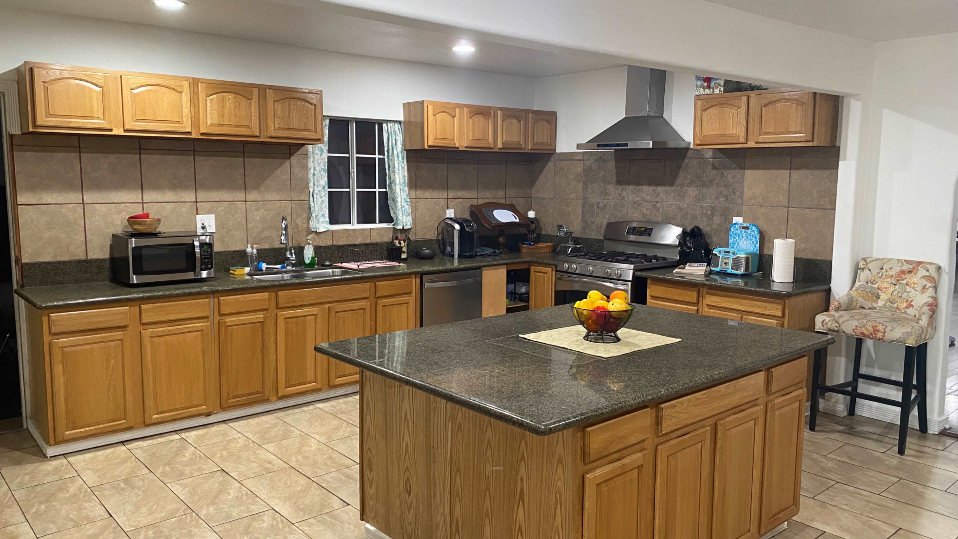 The kitchen features wooden cabinets, granite countertops, and stainless steel appliances, with a central island and a bowl of oranges on the countertop.