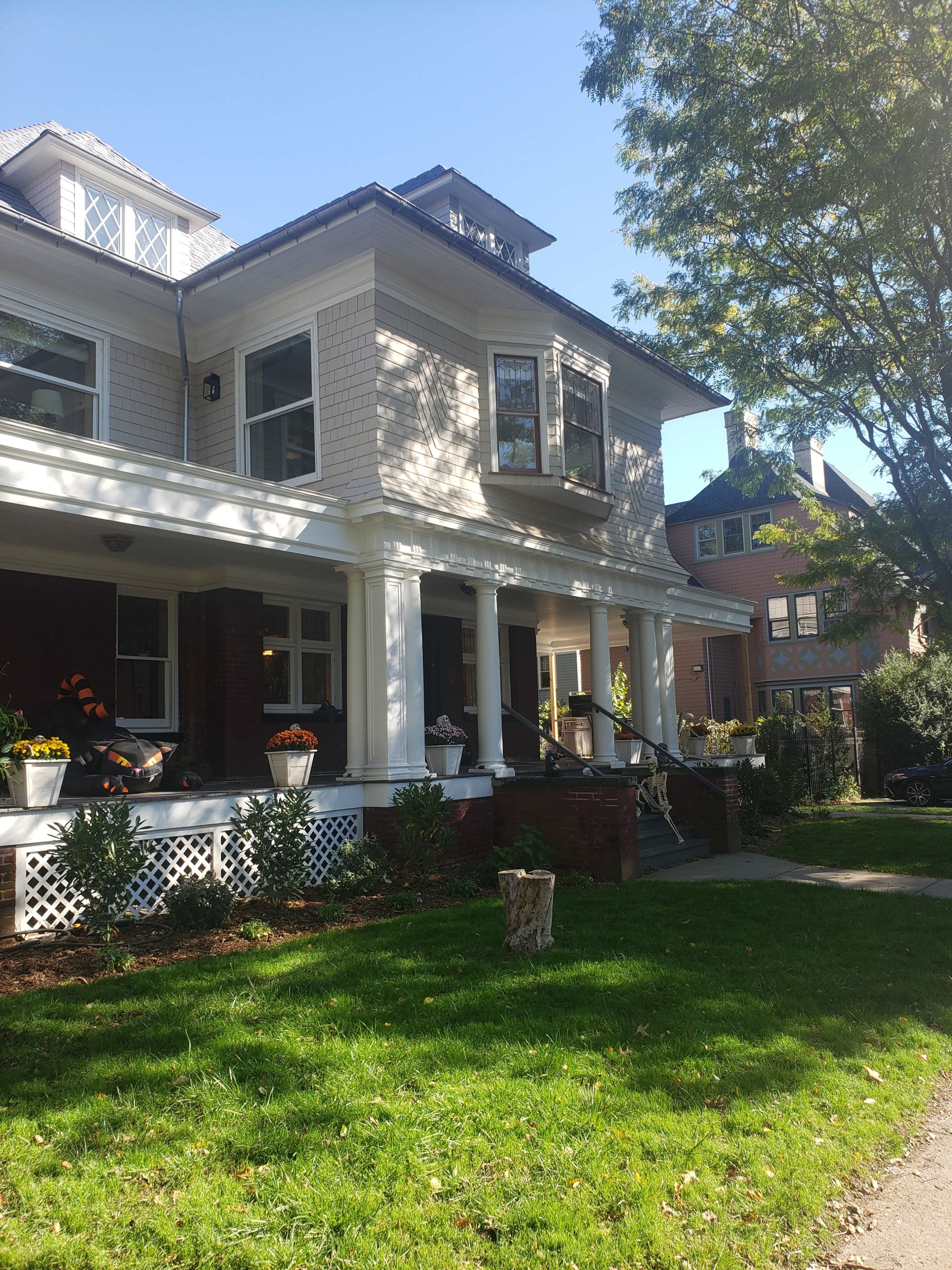 The image shows a two-story house with a large porch, surrounded by greenery and other residential buildings nearby.